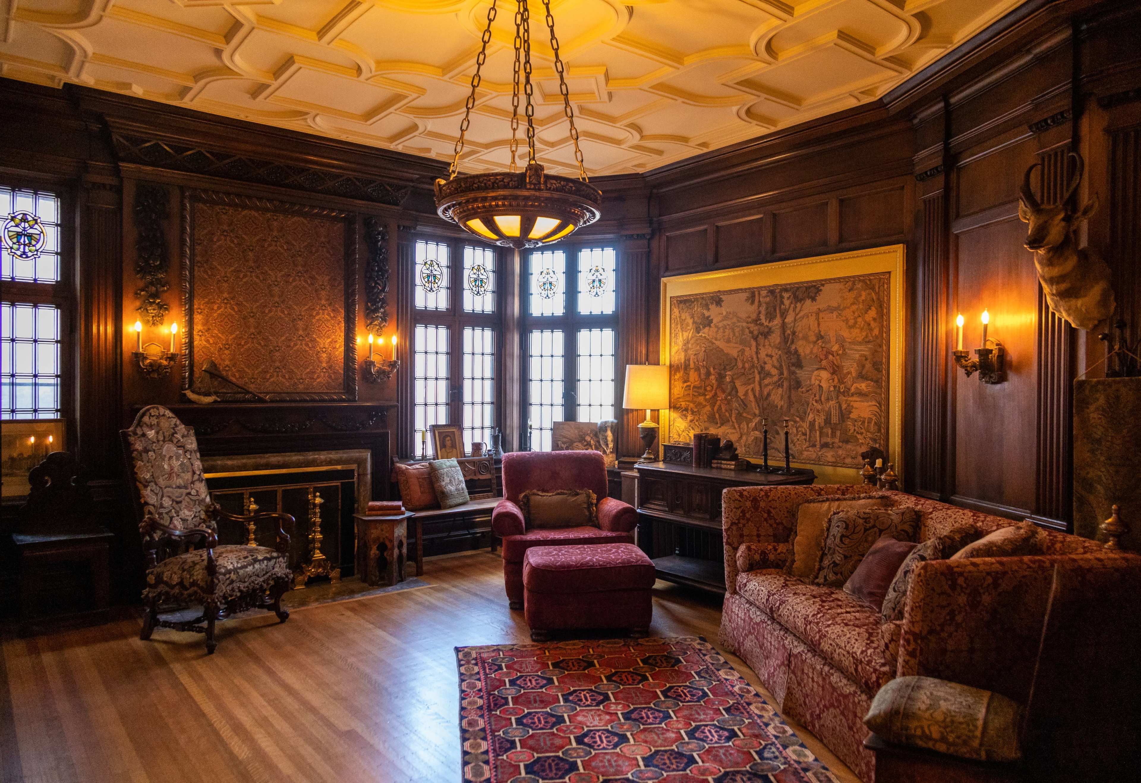 The image shows a elegantly furnished living room with wooden paneling, ornate light fixtures, and a large tapestry on the wall, centered around a fireplace.