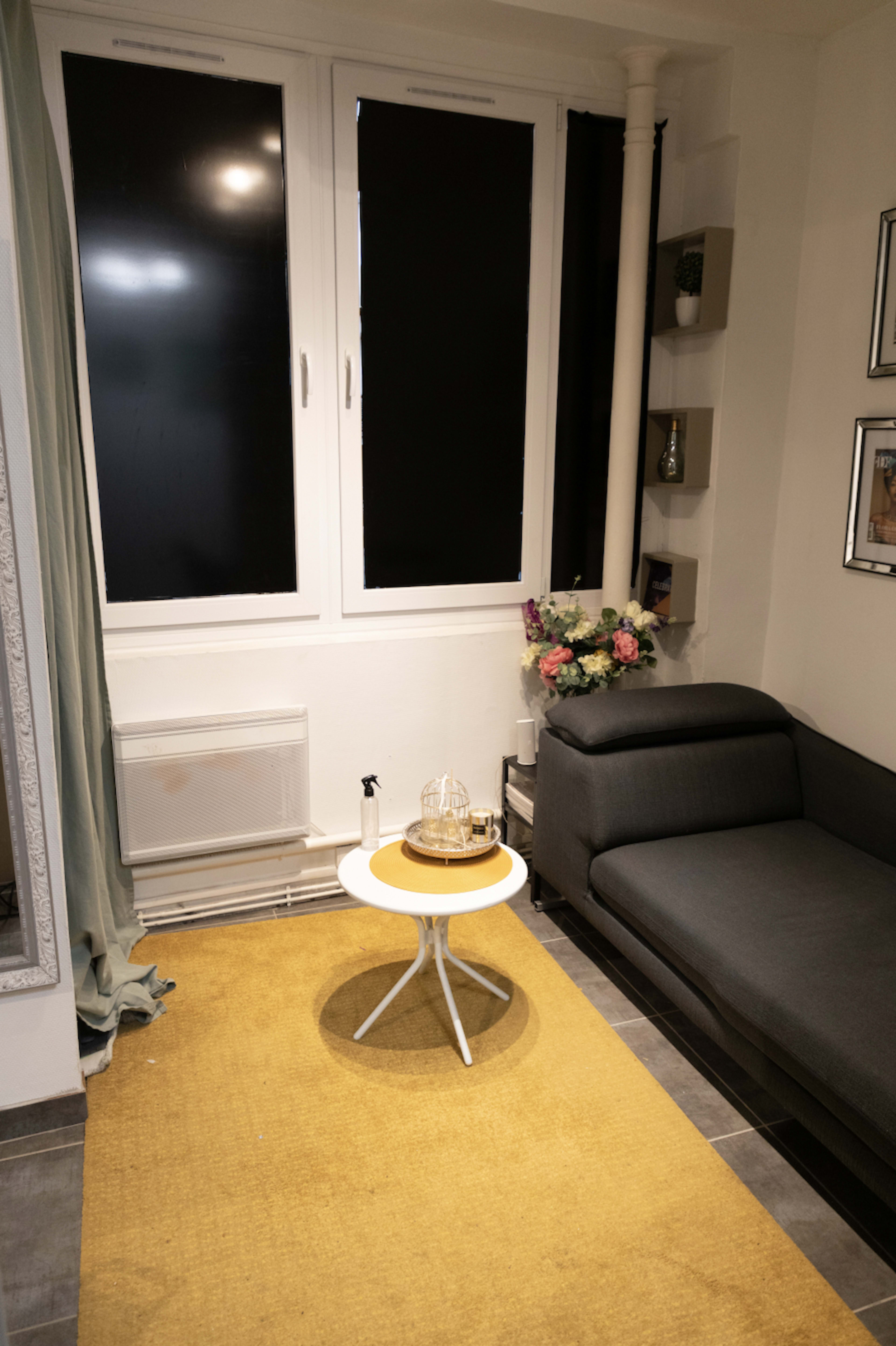 A gray sectional sofa sits next to a small round table on a yellow rug in a well-lit living room with a large window and decorative flowers.