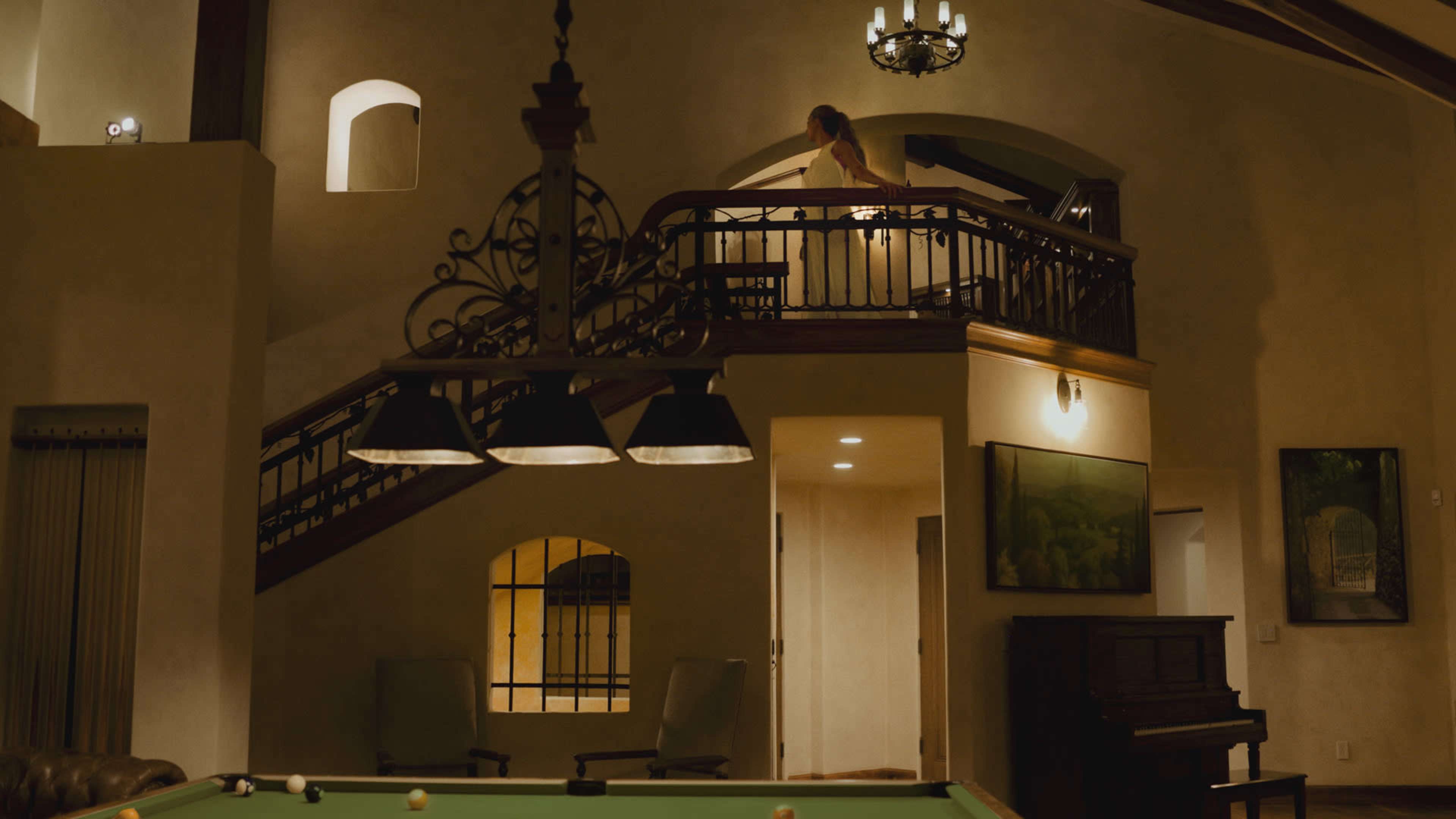 A person walks along a balcony overlooking a spacious interior with a pool table and a grand piano in a well-lit, elegantly designed room.