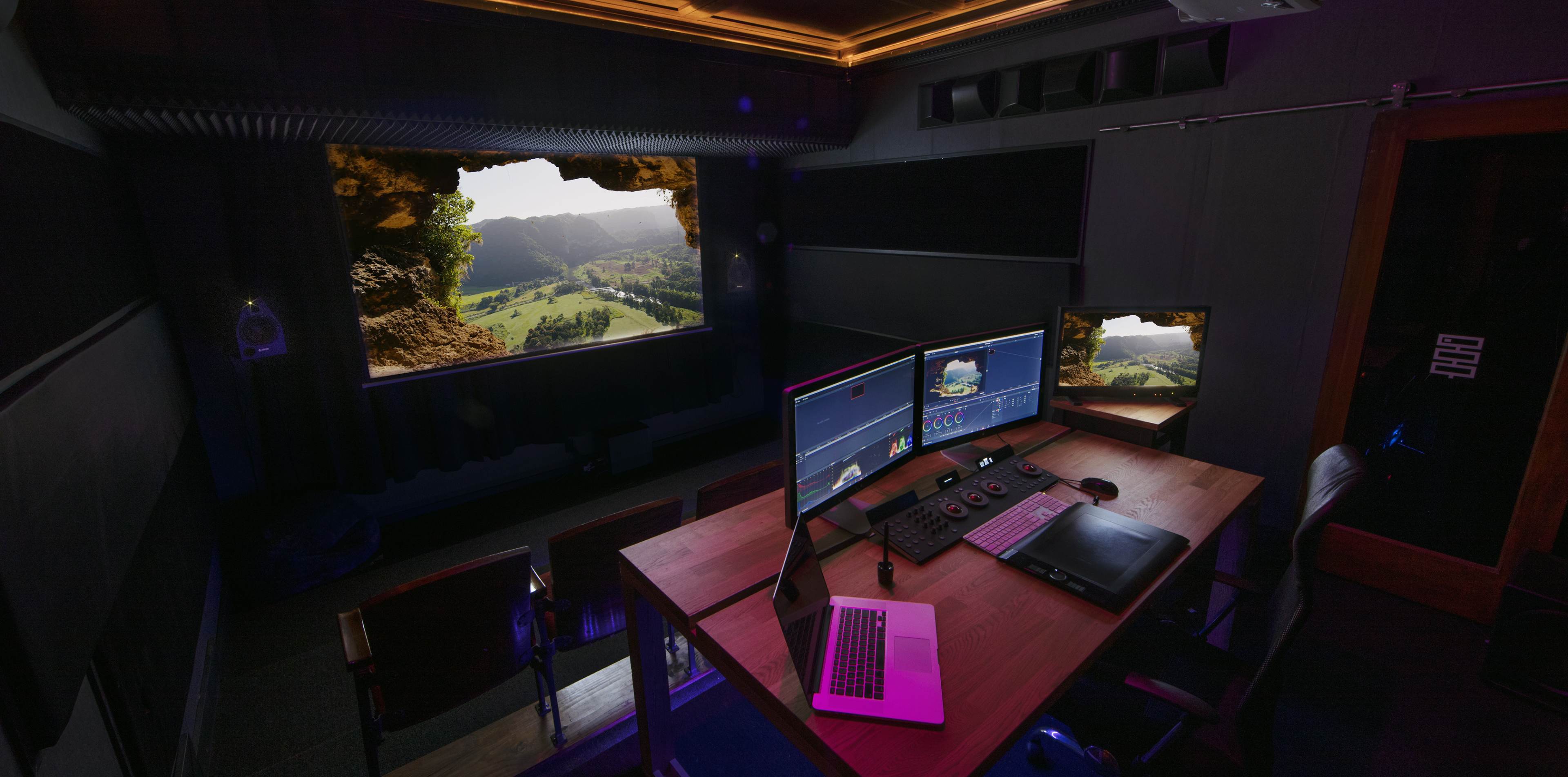The image shows a modern editing studio with two computer monitors and a laptop, featuring a view of a scenic landscape through a window-like opening in a stone wall.