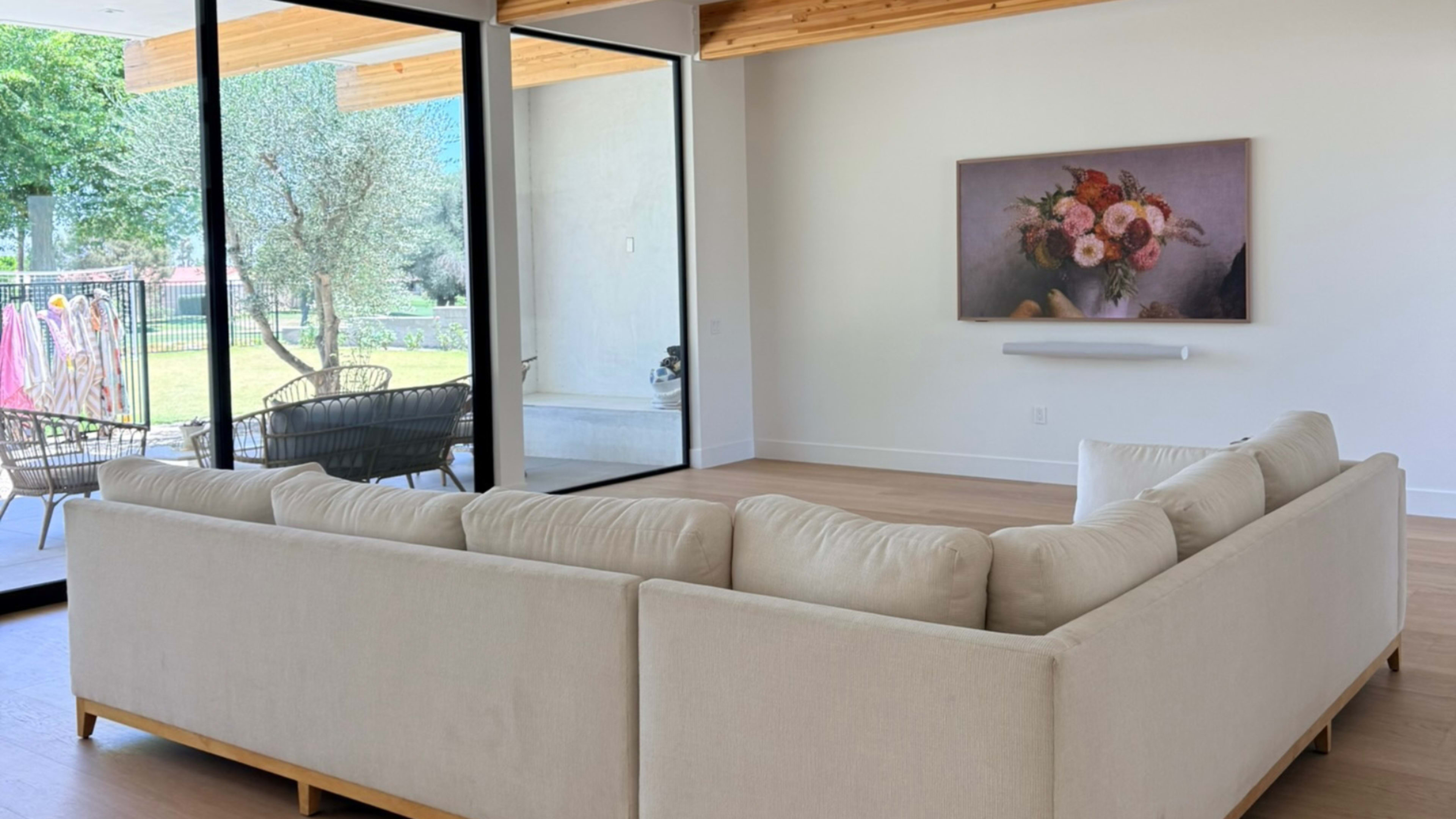 A large, light-colored sectional sofa faces a framed floral painting on the wall in a bright living room with large glass doors opening to a garden.