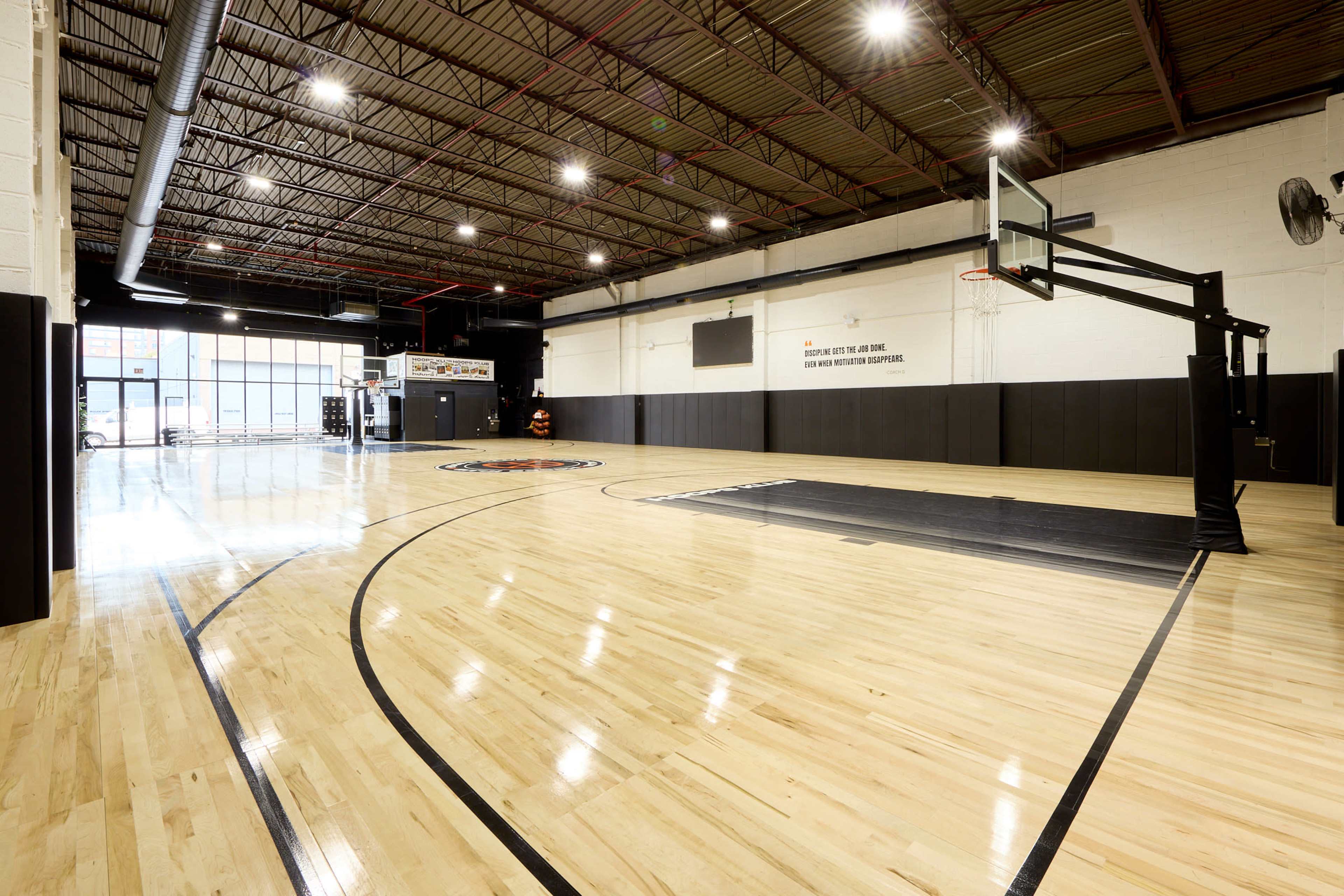 The image shows a spacious indoor basketball court with polished wooden flooring, a hoop at one end, and large windows allowing light to enter.