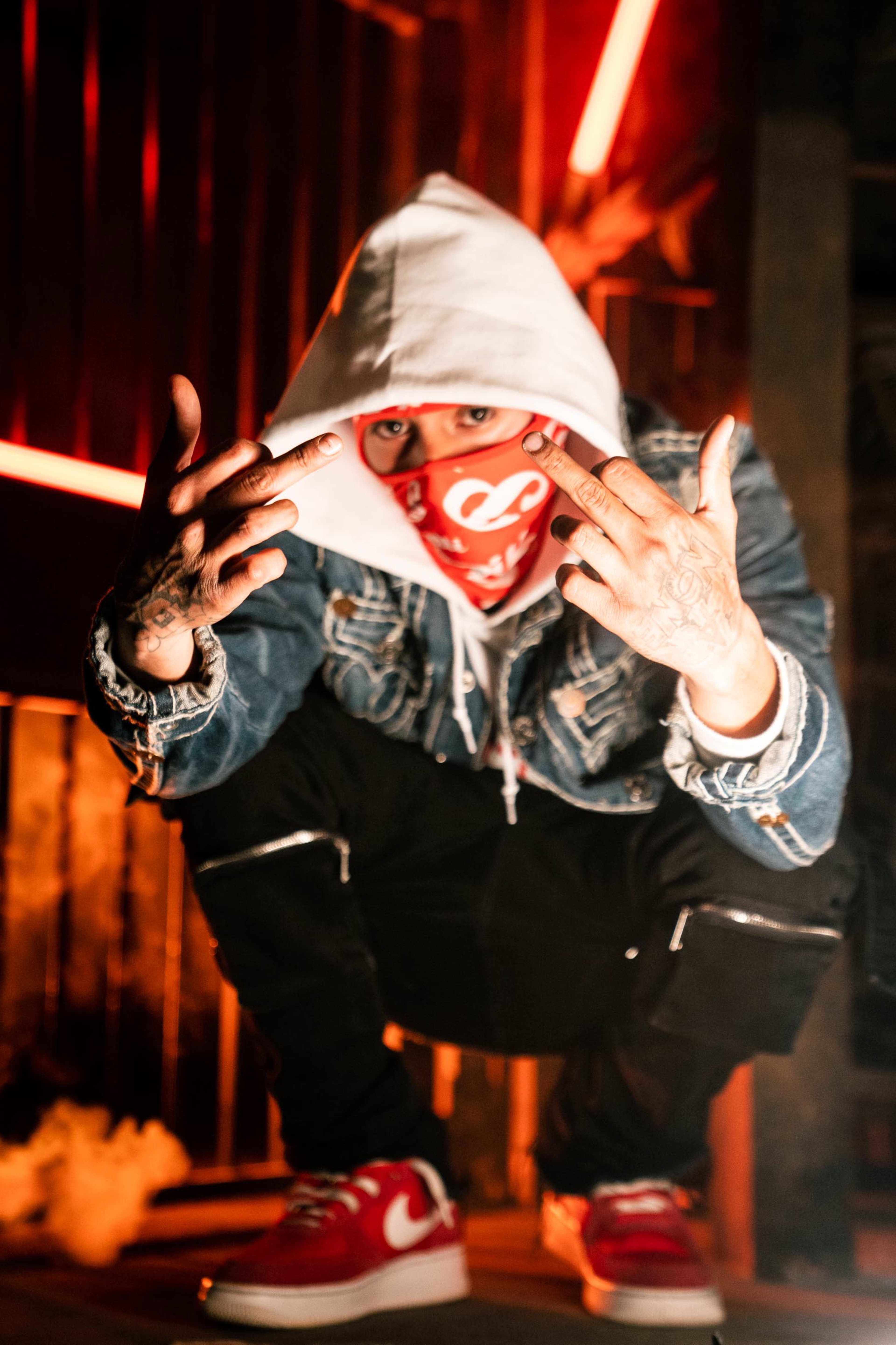 A person wearing a hooded sweatshirt and bandana poses with their middle fingers raised while squatting against a backdrop of dim lighting and smoke.