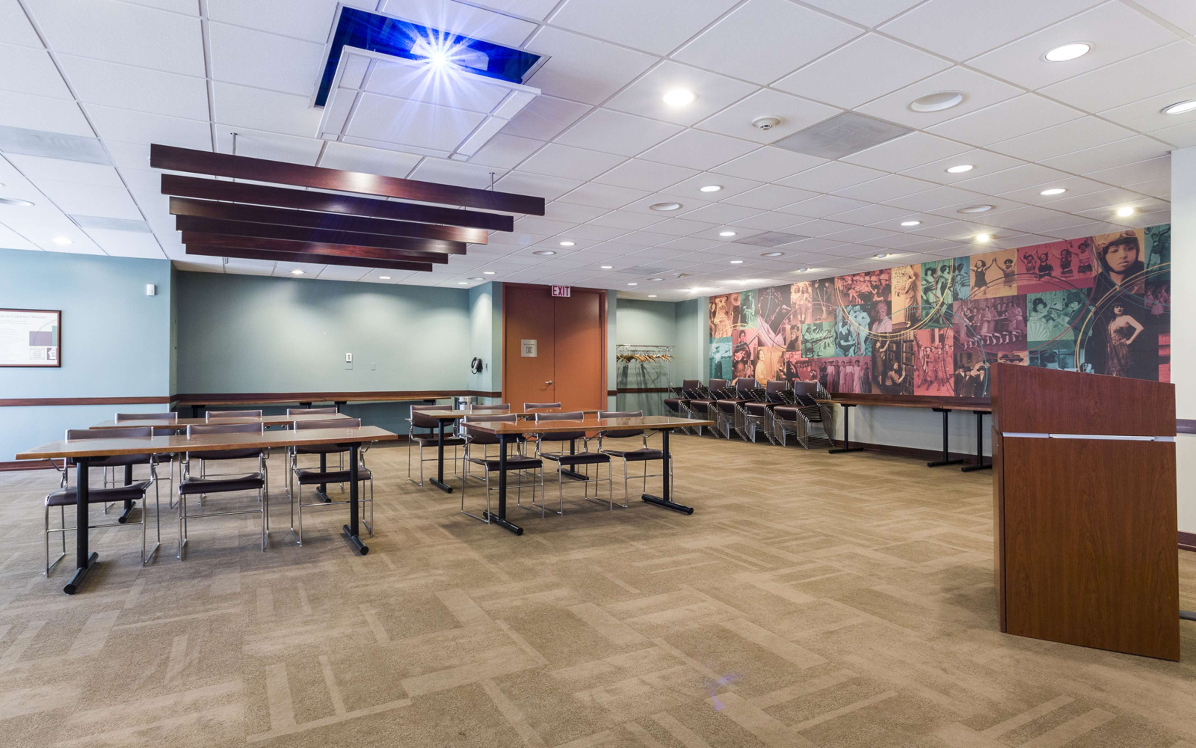 A spacious room with tables and chairs arranged for a meeting or event, featuring a colorful mural on one wall and a reception area.