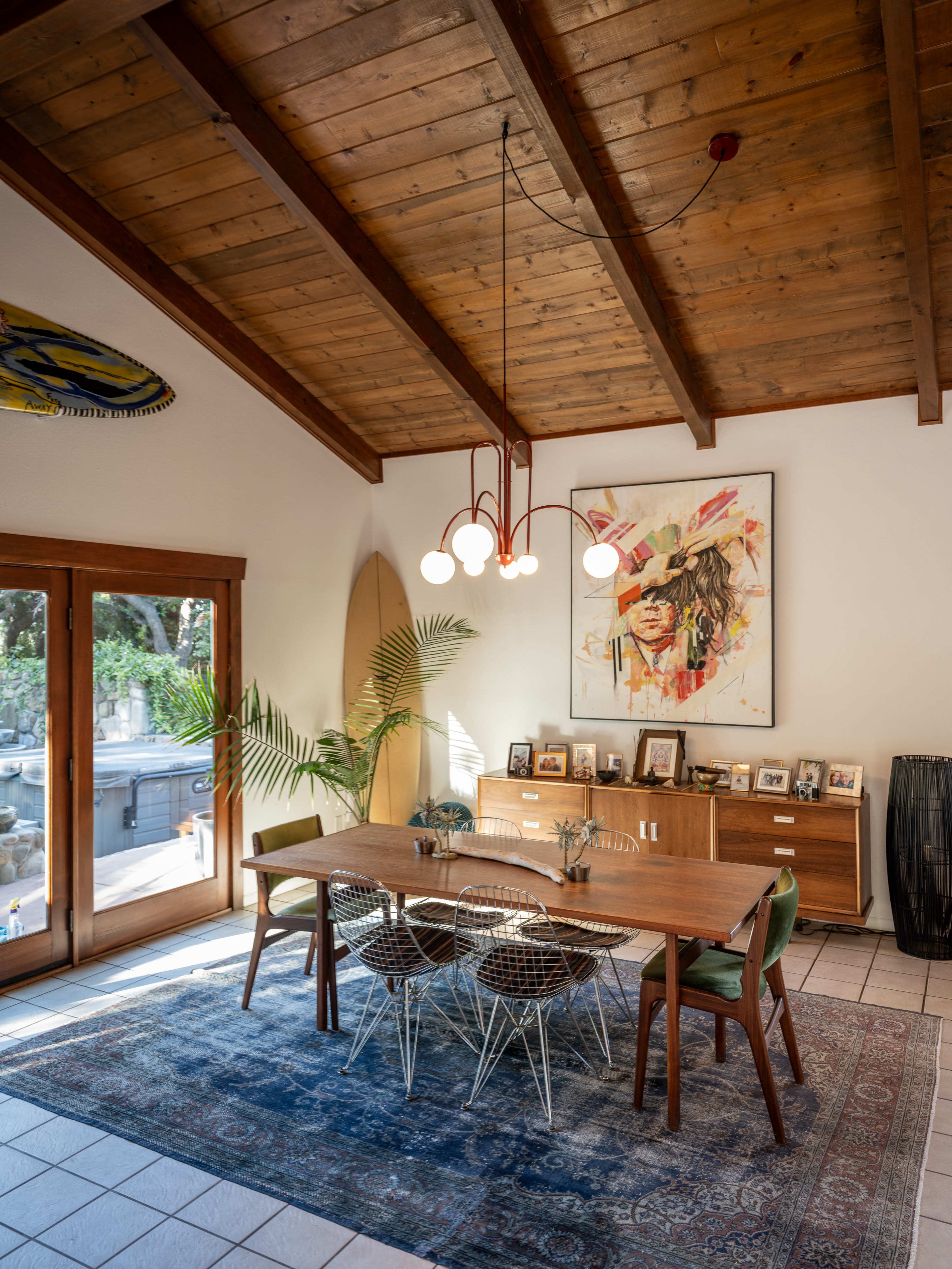 A dining area features a wooden table surrounded by chairs, a large artwork on the wall, and a surfboard leaning against the side, with a backdrop of large windows.
