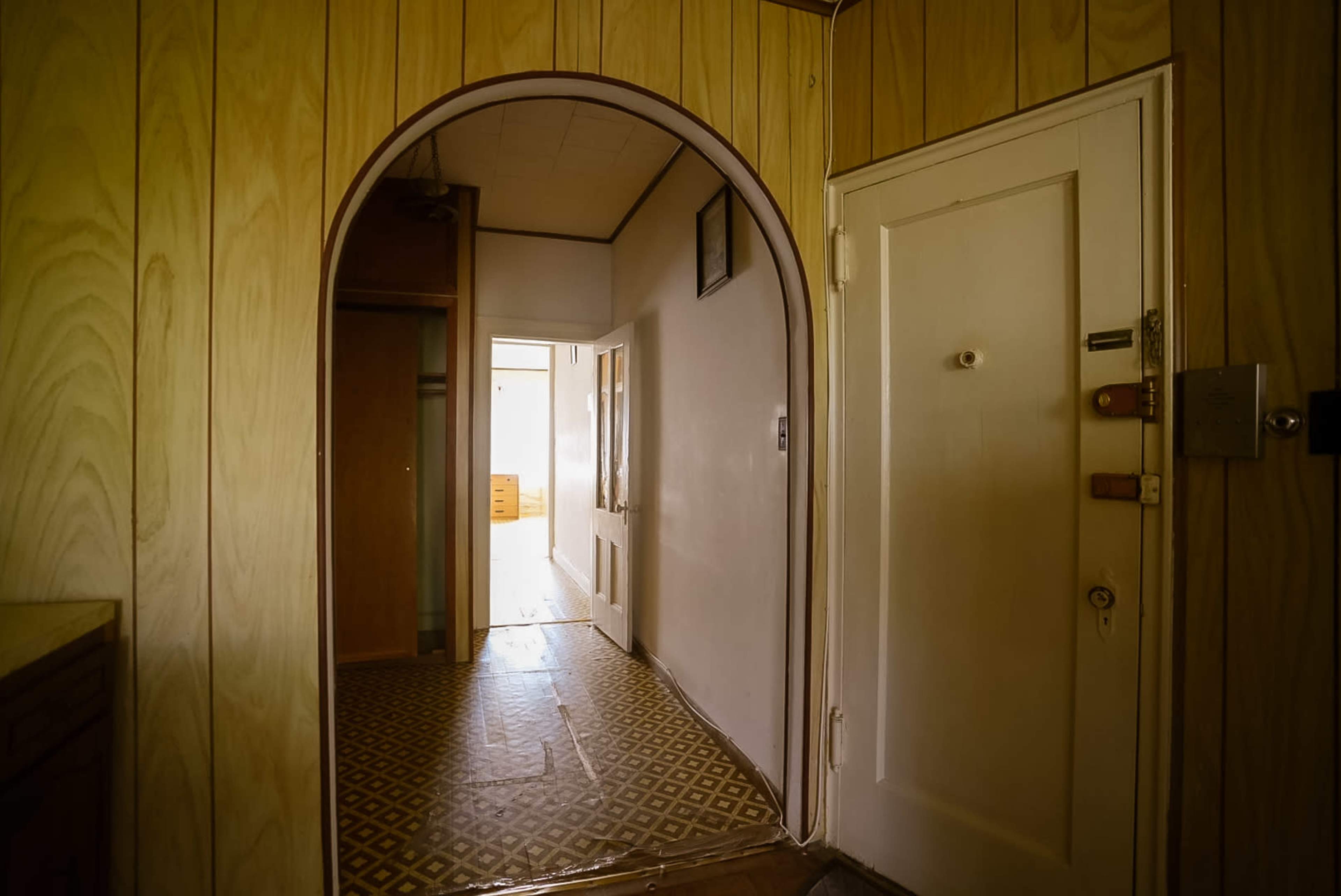 A narrow hallway with wooden paneling leads to an archway and a door, showcasing light from an adjacent room.