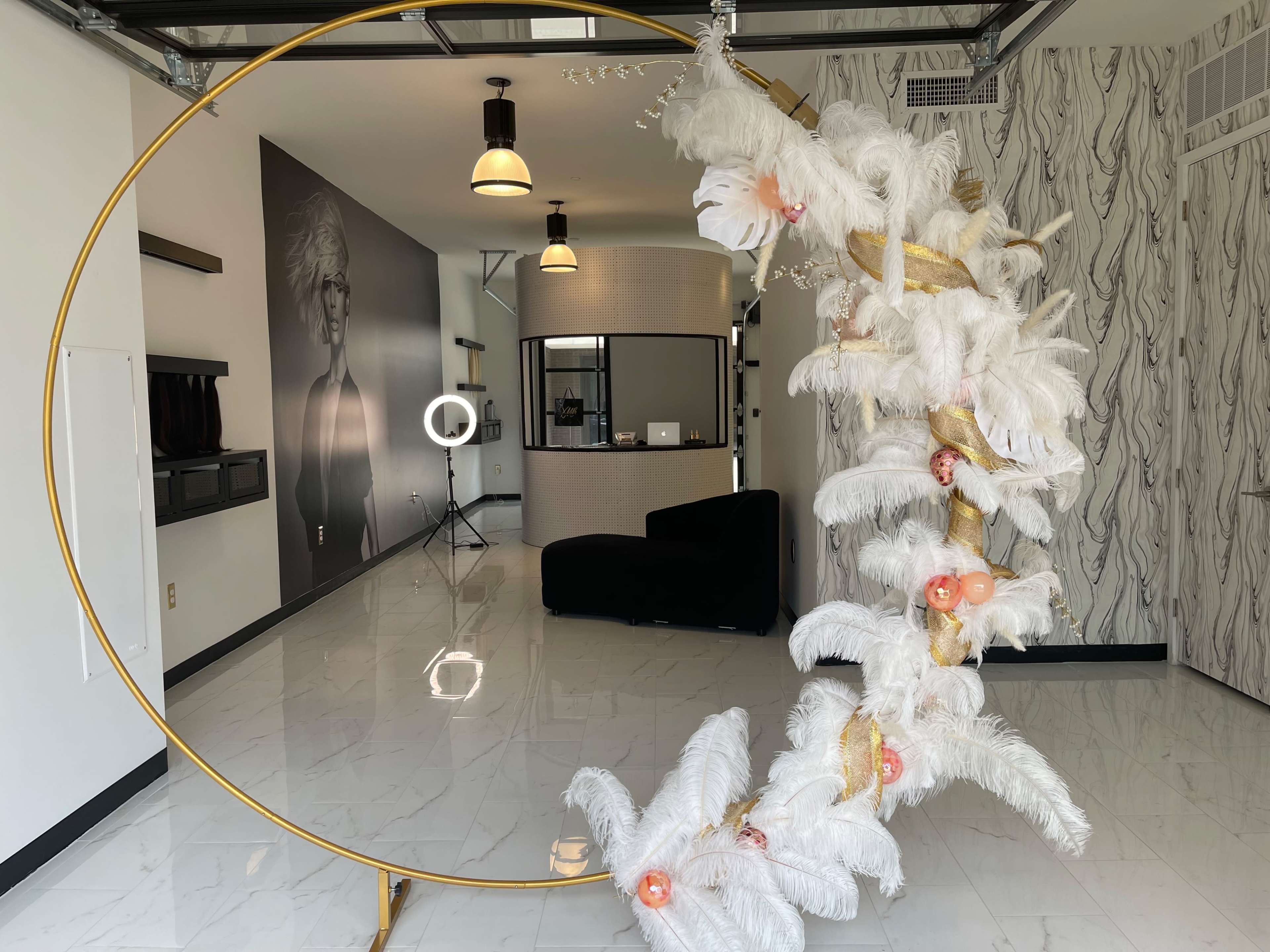 A decorative circular structure made of gold and adorned with white feathers and floral elements stands in a modern salon with a stylish reception area in the background.