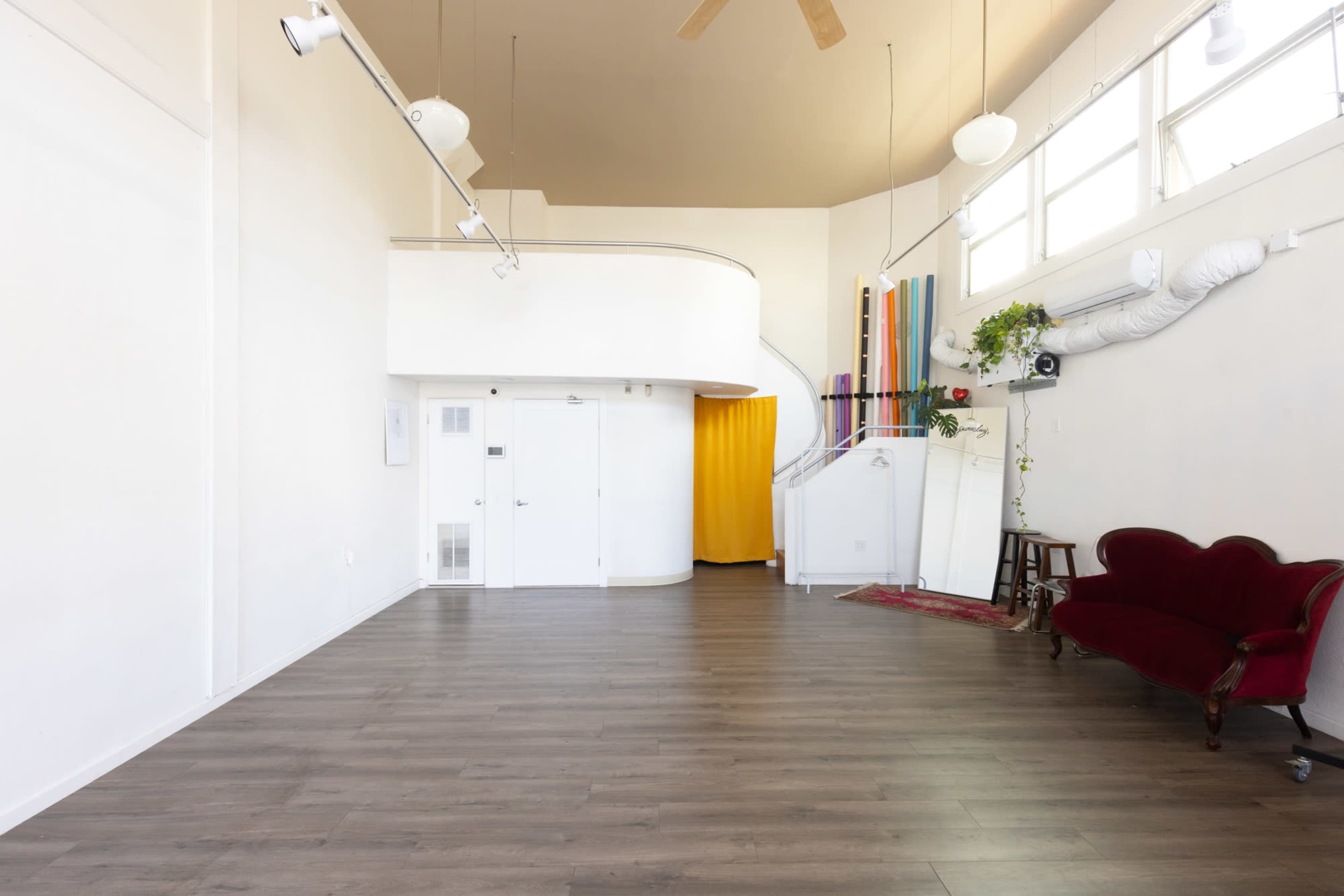 A spacious room features light-colored walls, wooden flooring, and a plush red couch, with a yellow curtain at the entrance of a side room.