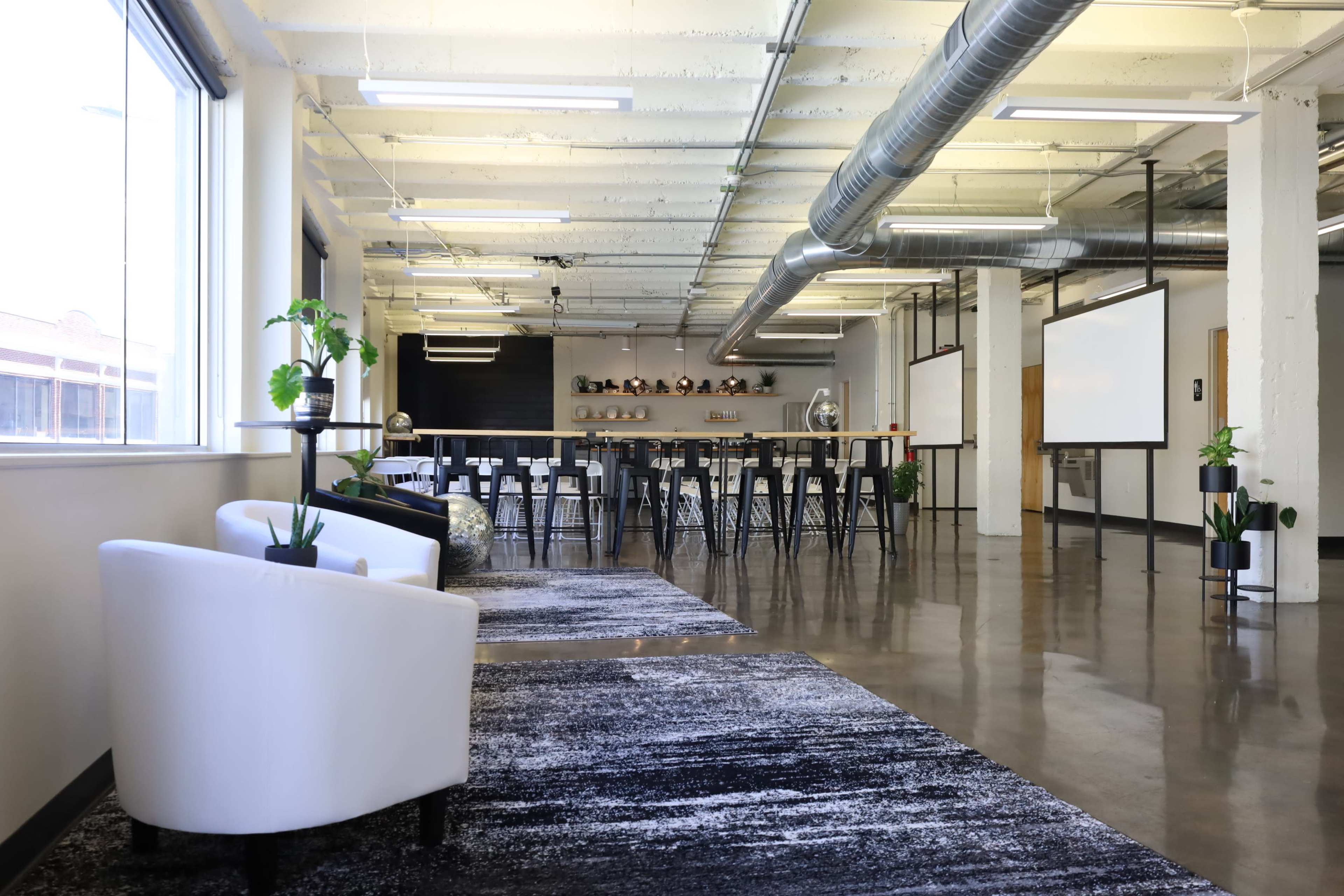 A large, open workspace with modern furnishings, including white chairs, a row of tall tables, and greenery, under bright overhead lighting.