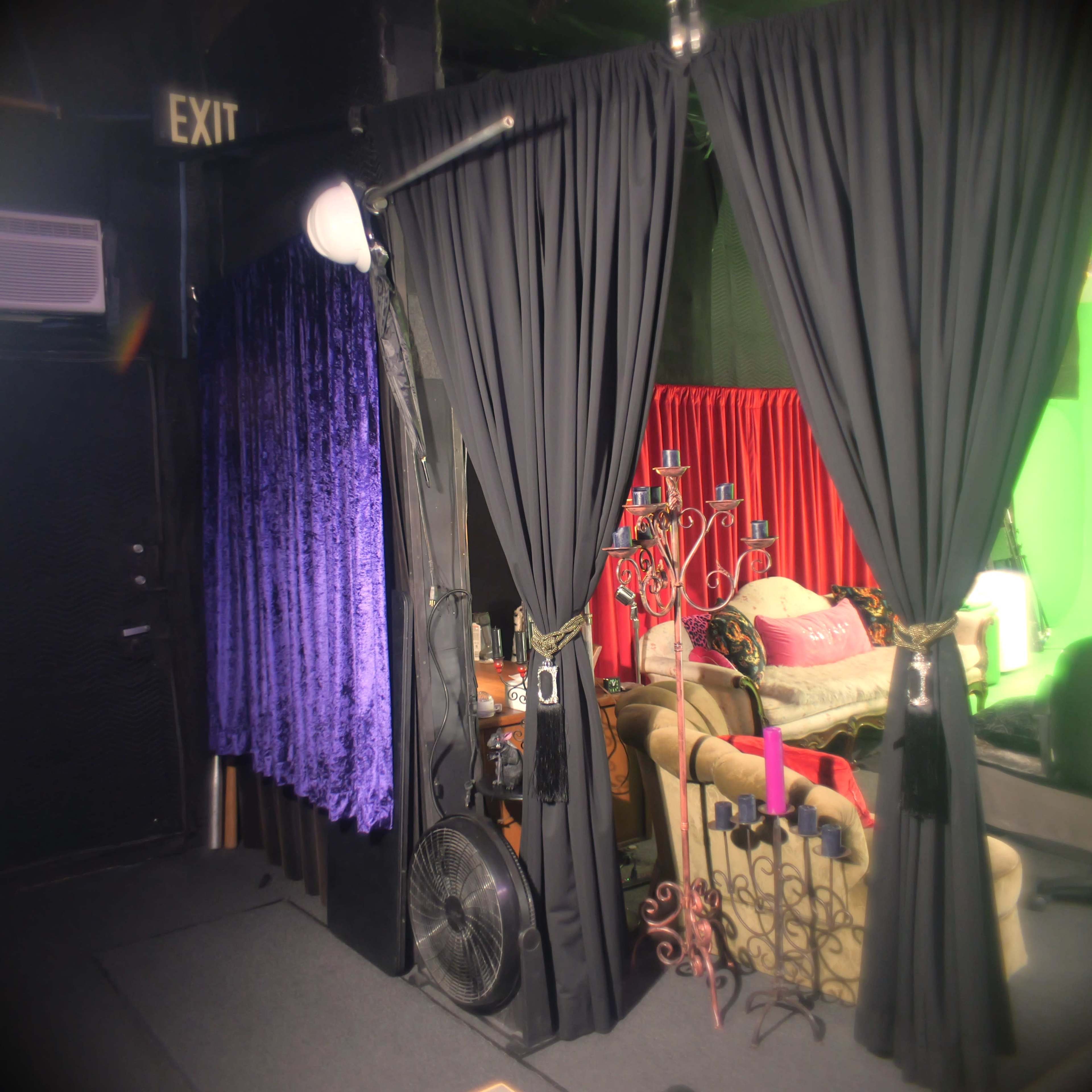The image shows a dimly lit space with black, purple, and red curtains separating different areas, along with a fan and some furniture visible in the background.