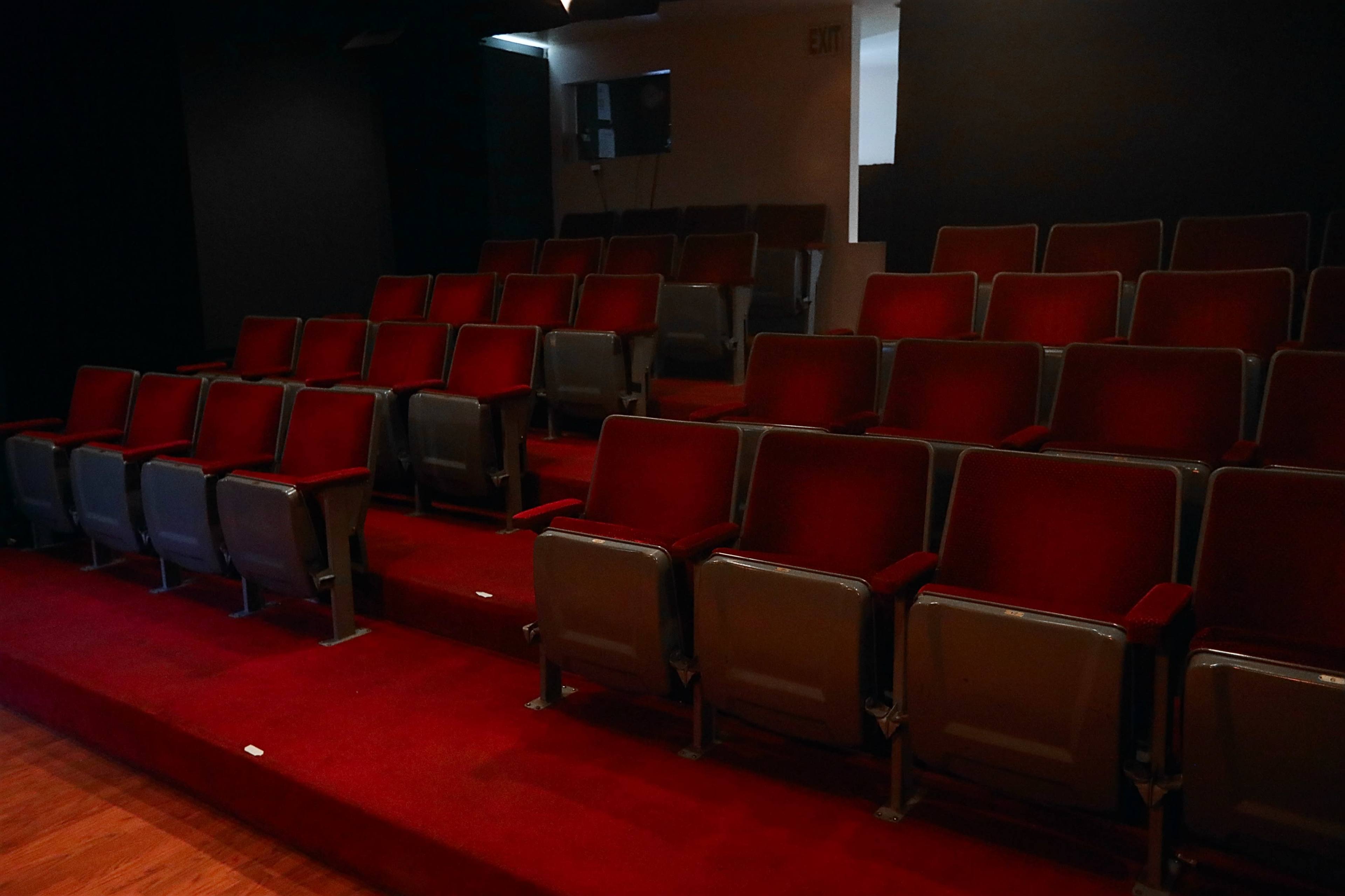 The image shows a row of empty red theater seats arranged on a carpeted step in a dimly lit auditorium.
