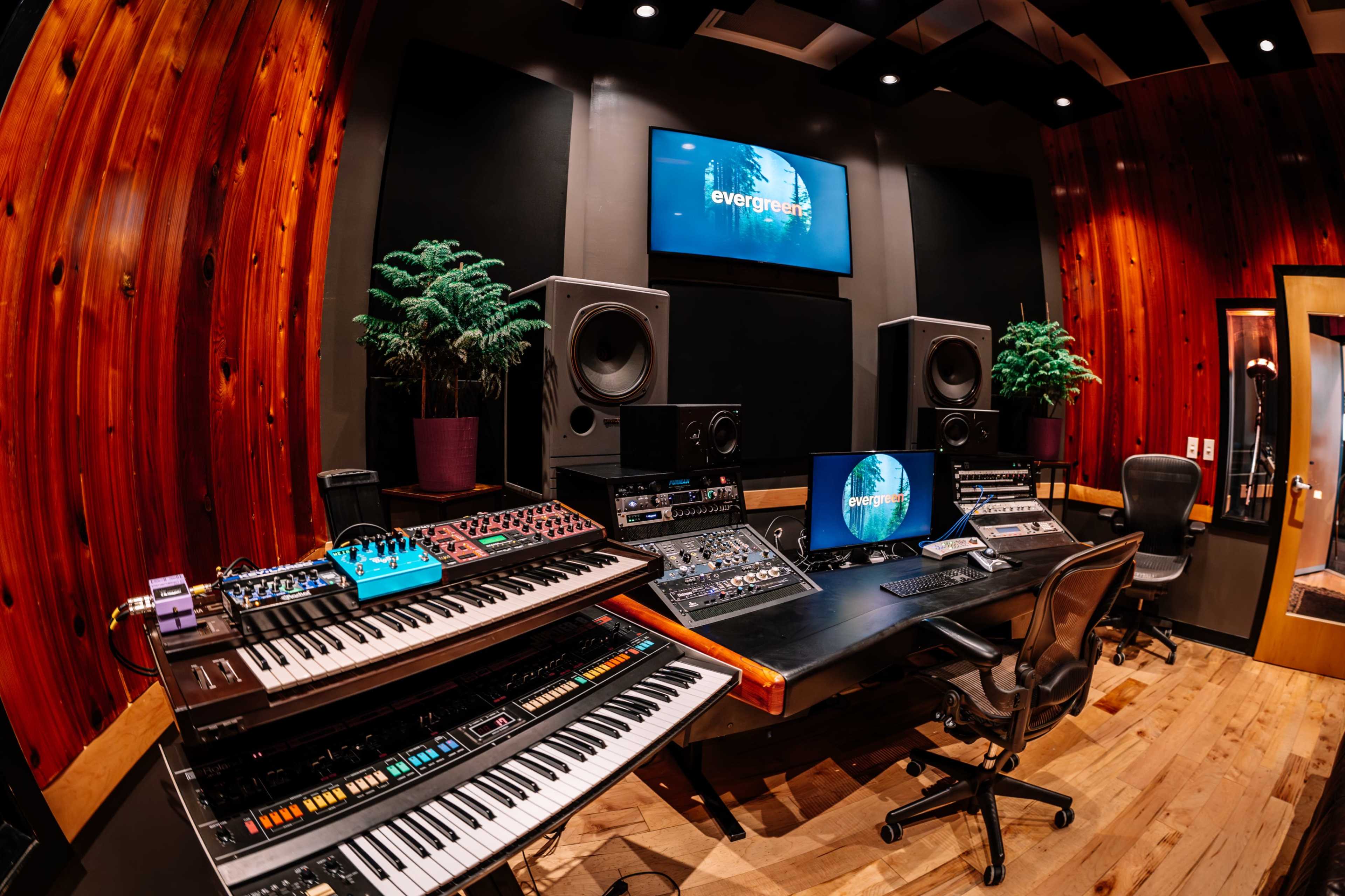The image shows a music recording studio with a variety of synthesizers, mixing equipment, large speakers, and a desk featuring a computer and display in a wood-paneled room.