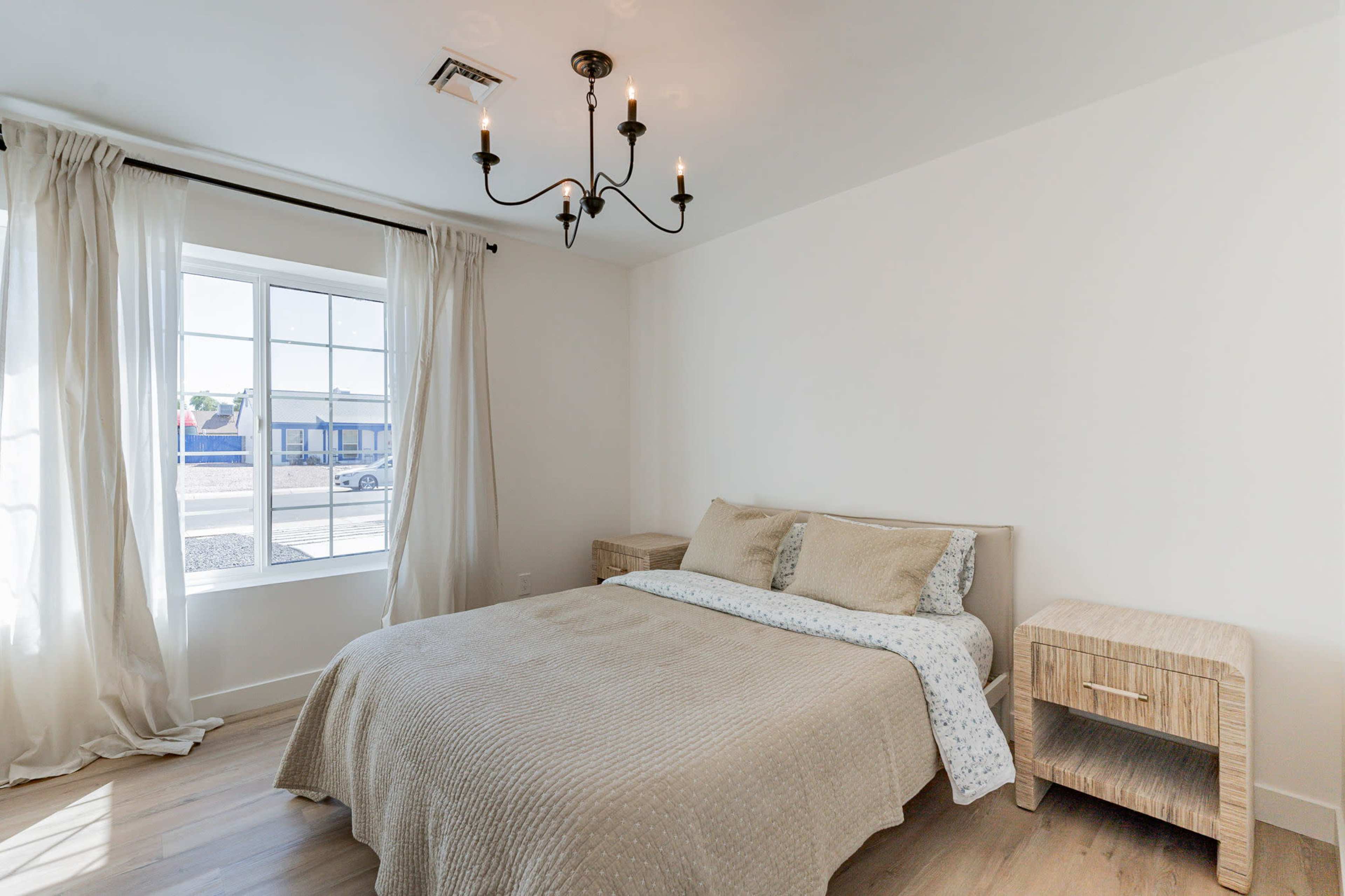 The image shows a simple bedroom with a bed, two bedside tables, a chandelier, and a large window allowing natural light to enter.