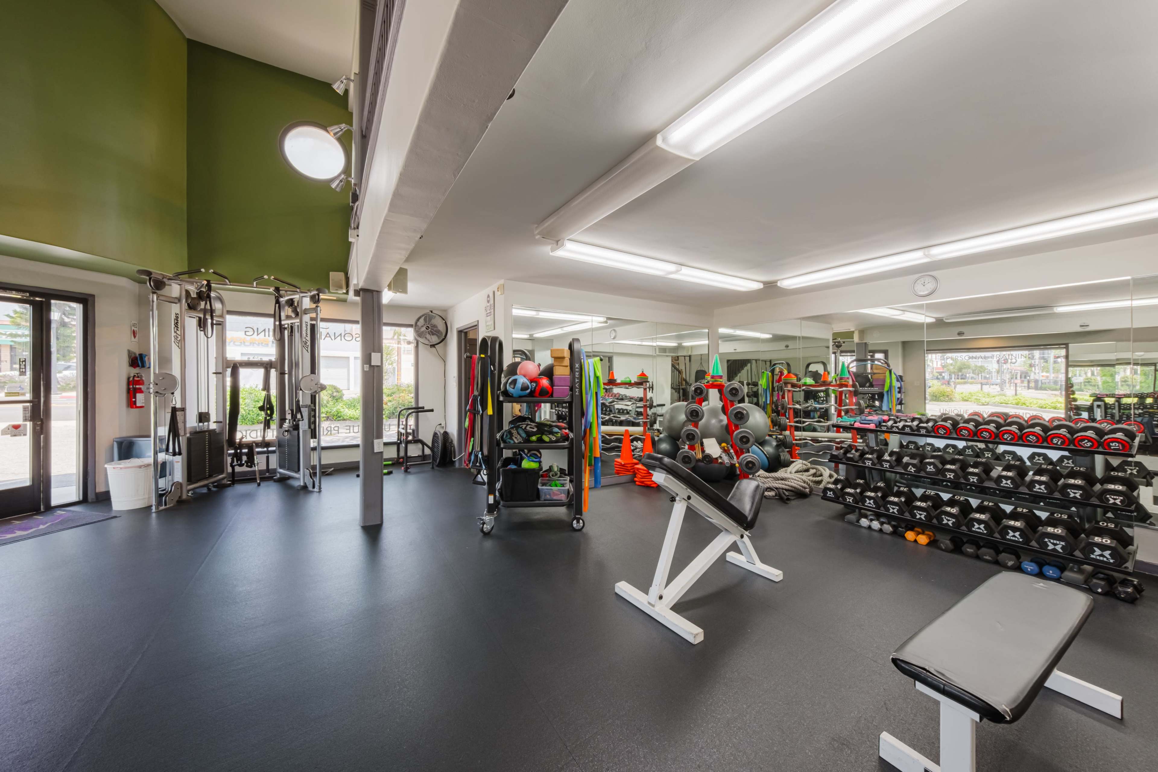 A fitness gym with various weightlifting equipment, including dumbbells on racks and benches, and bright lighting throughout the space.
