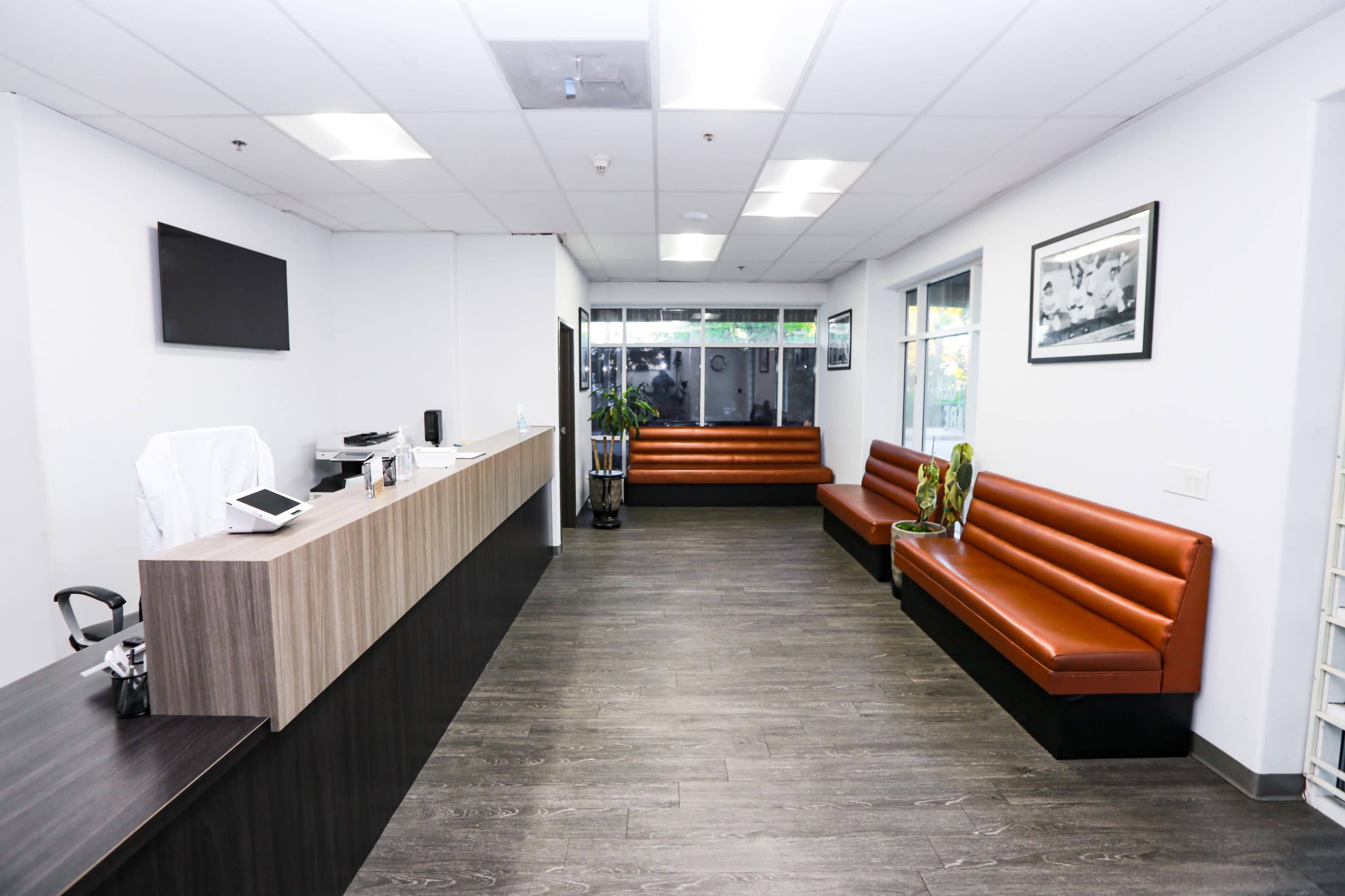 The image shows a modern waiting area with brown benches, a reception desk, and large windows letting in natural light.