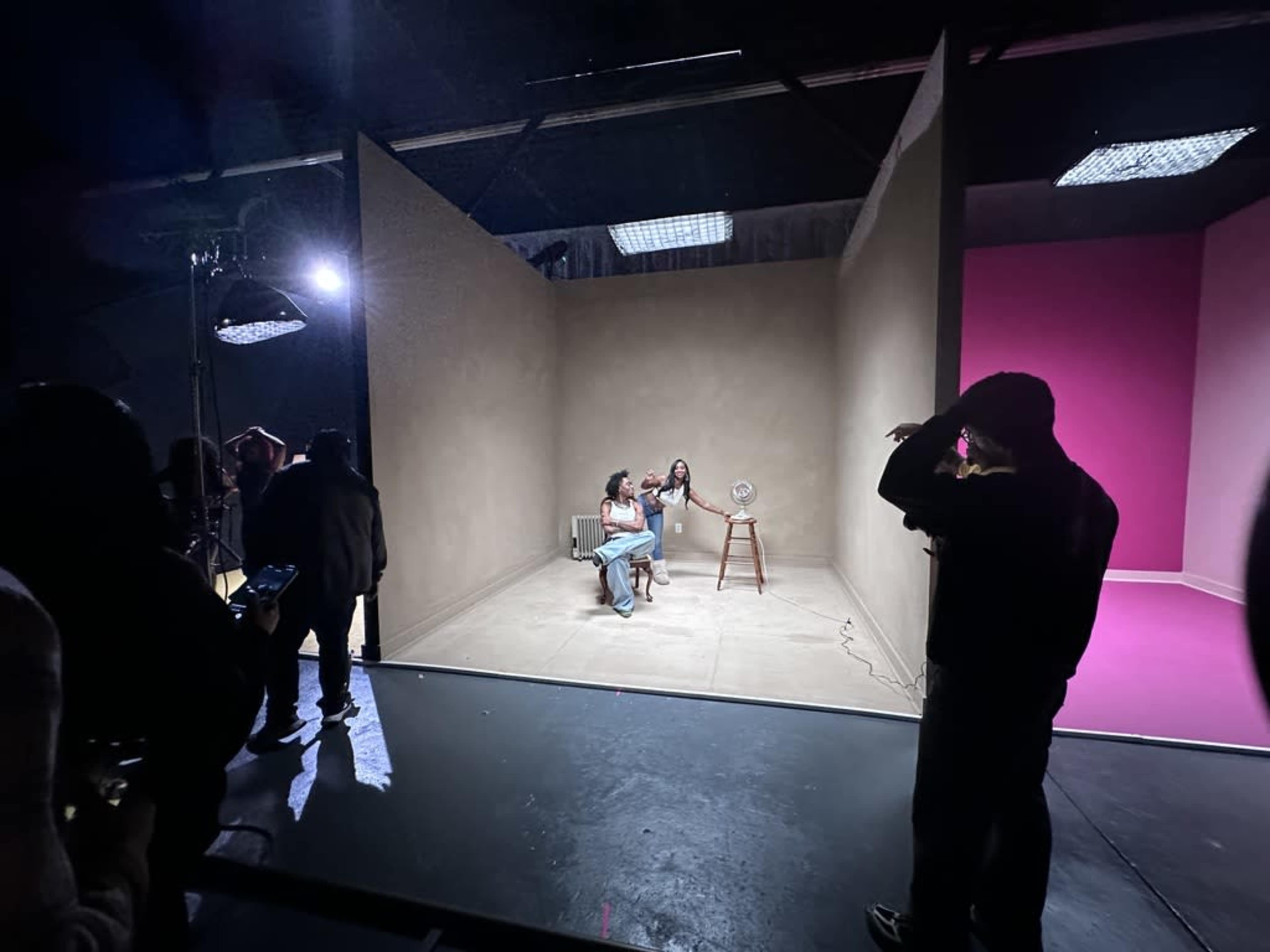 Two women are seated on a chair in a studio with contrasting backgrounds: one wall is beige and the other is pink, while people observe from the outside.