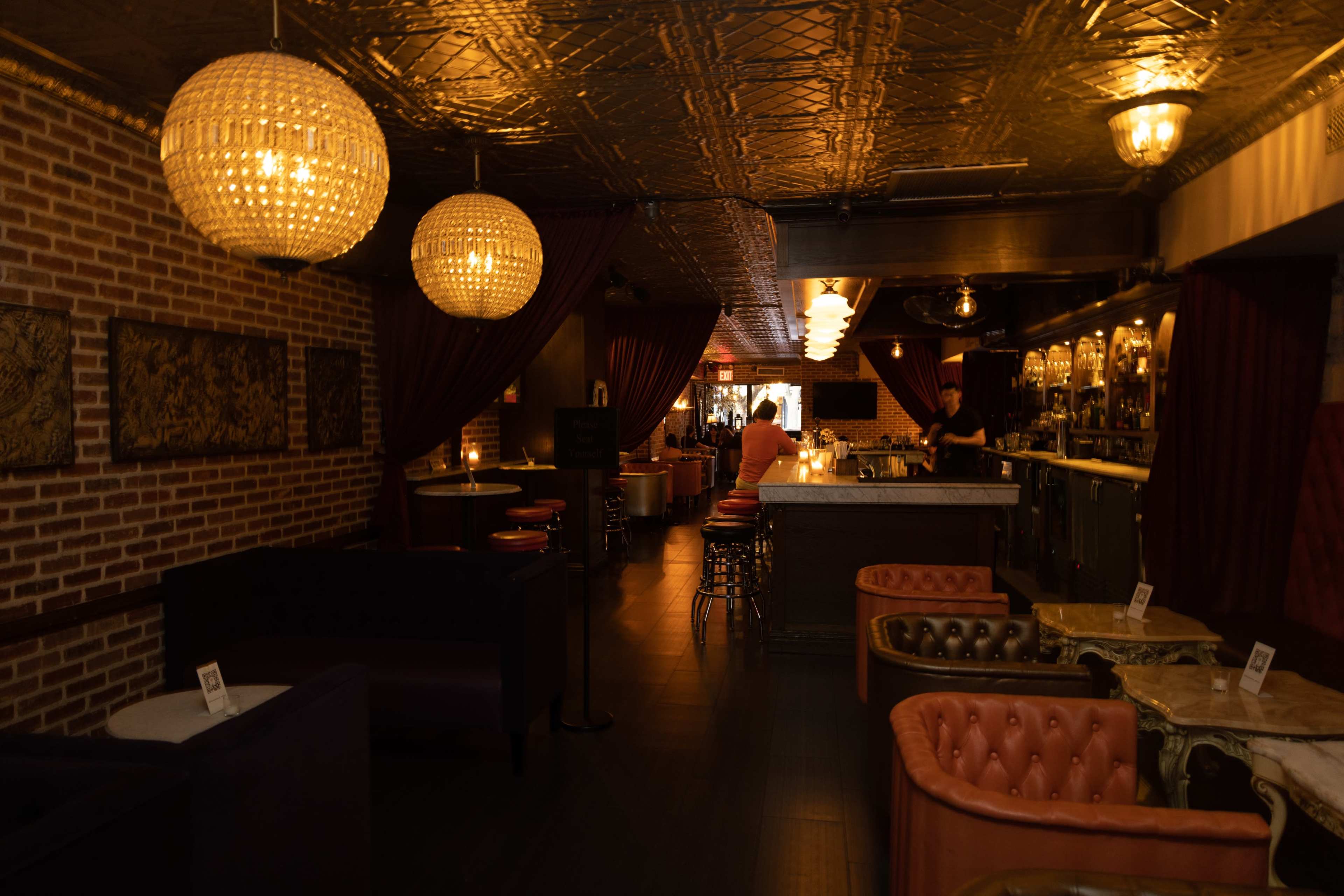 A dimly lit bar interior features brick walls, plush seating, and ornate chandeliers hanging from the ceiling.