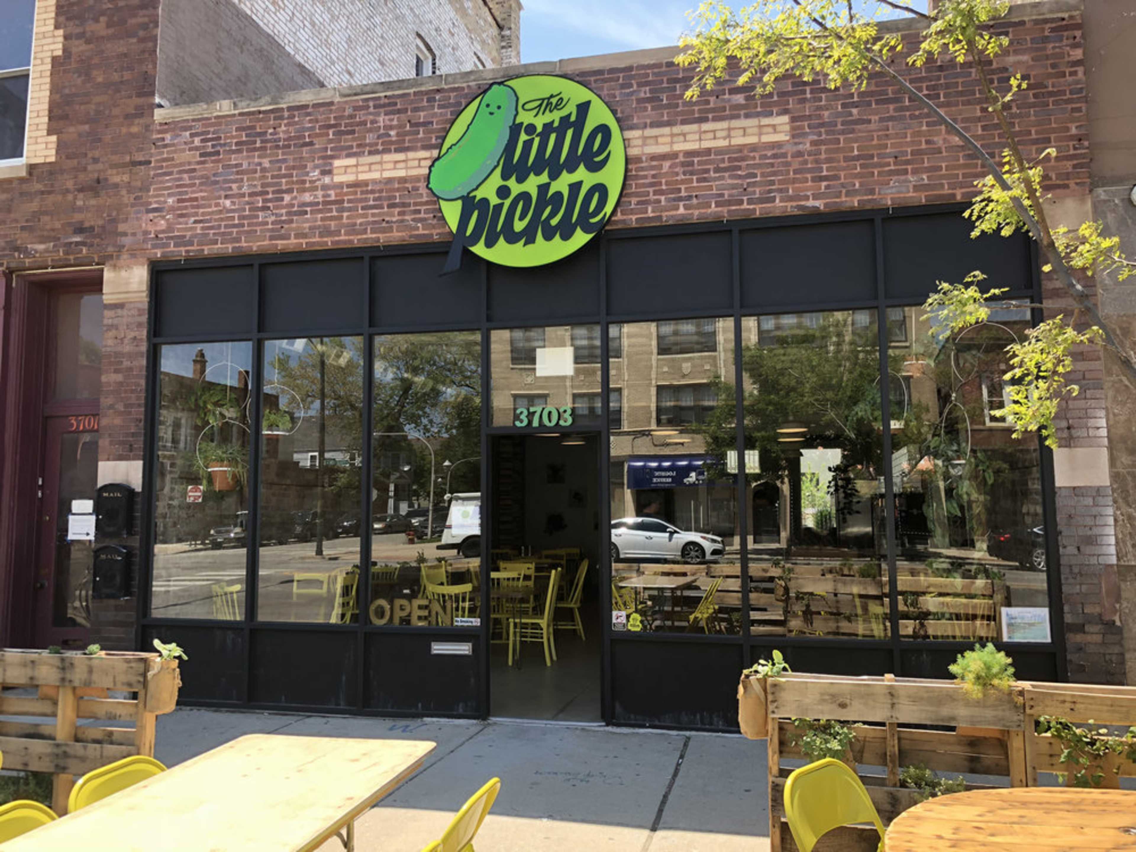 The storefront of a restaurant called "The Little Pickle," featuring large windows and outdoor seating with yellow chairs.