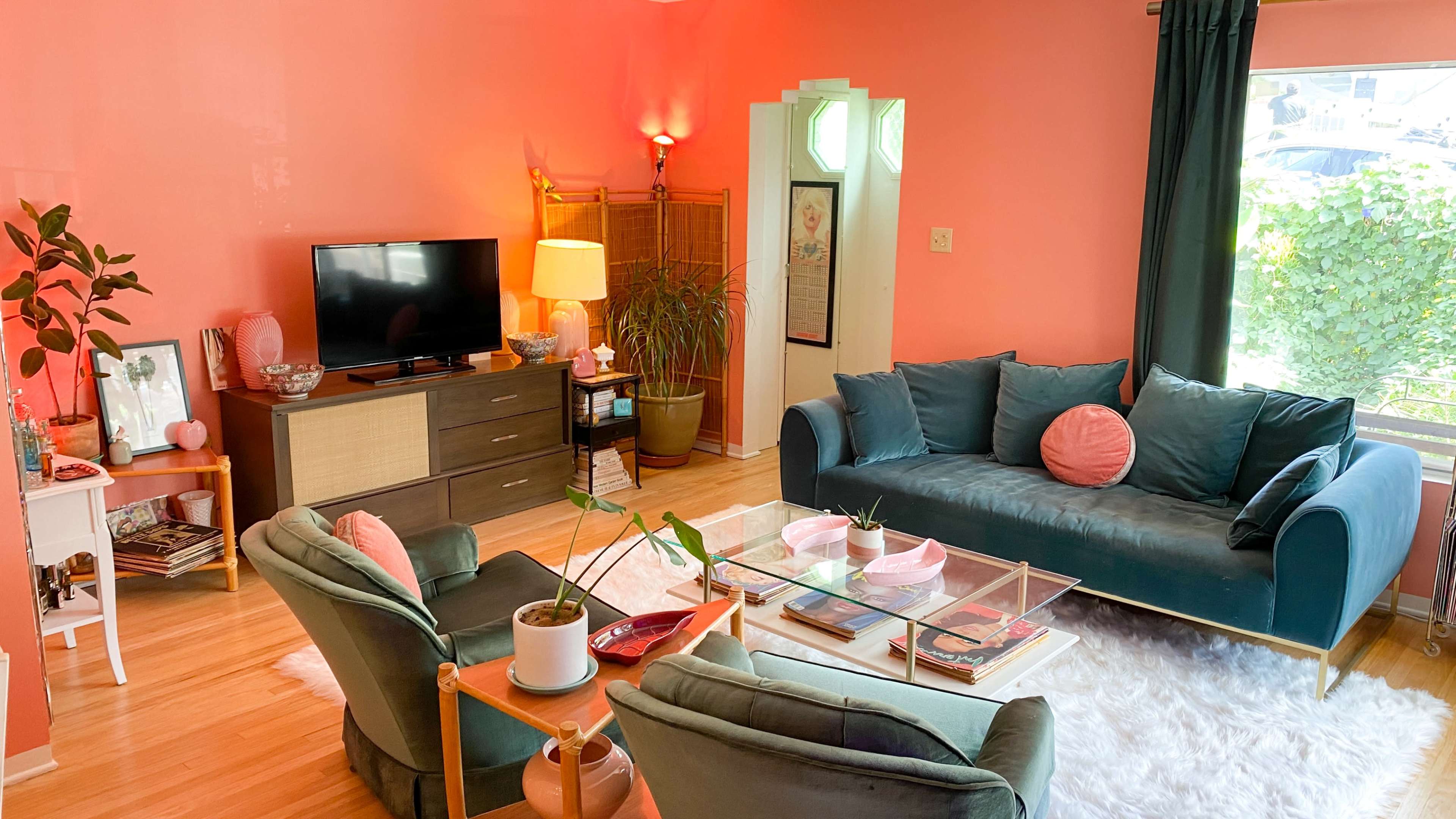 The living room features a coral-painted wall, a television on a wooden stand, two teal sofas with pink cushions, and a variety of plants.