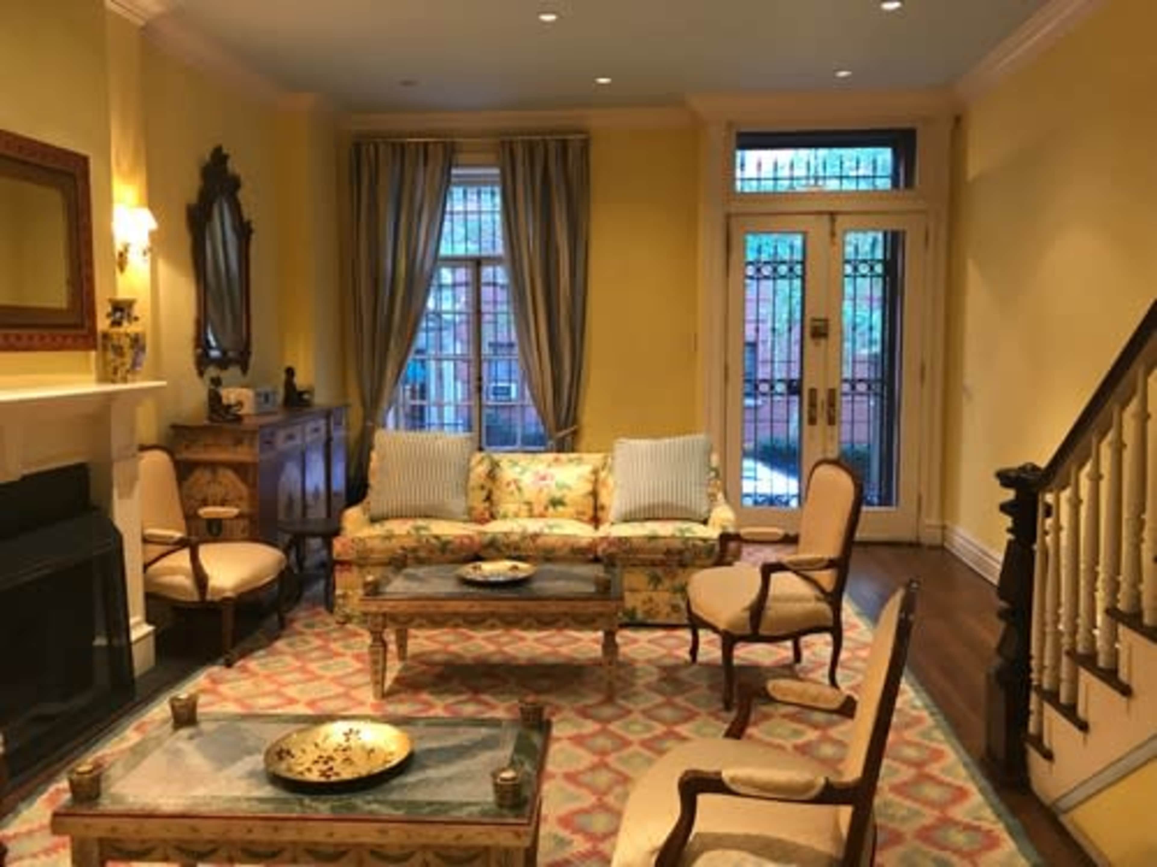 The room features a seating area with floral-patterned furniture, a coffee table, and a decorative rug, alongside a staircase and large windows.