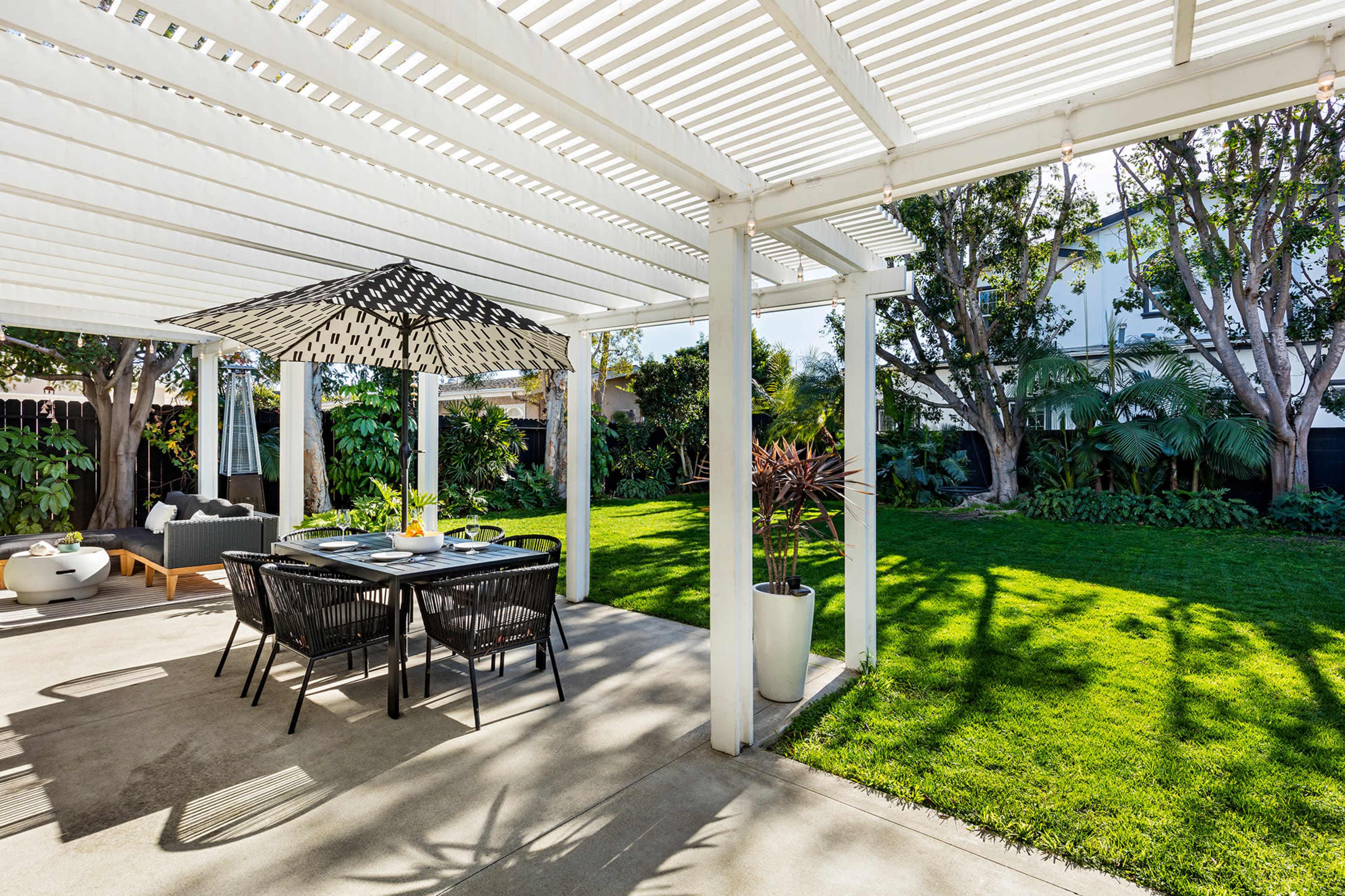 A covered outdoor dining area features a table with black chairs and an umbrella, surrounded by a well-maintained lawn and lush greenery.