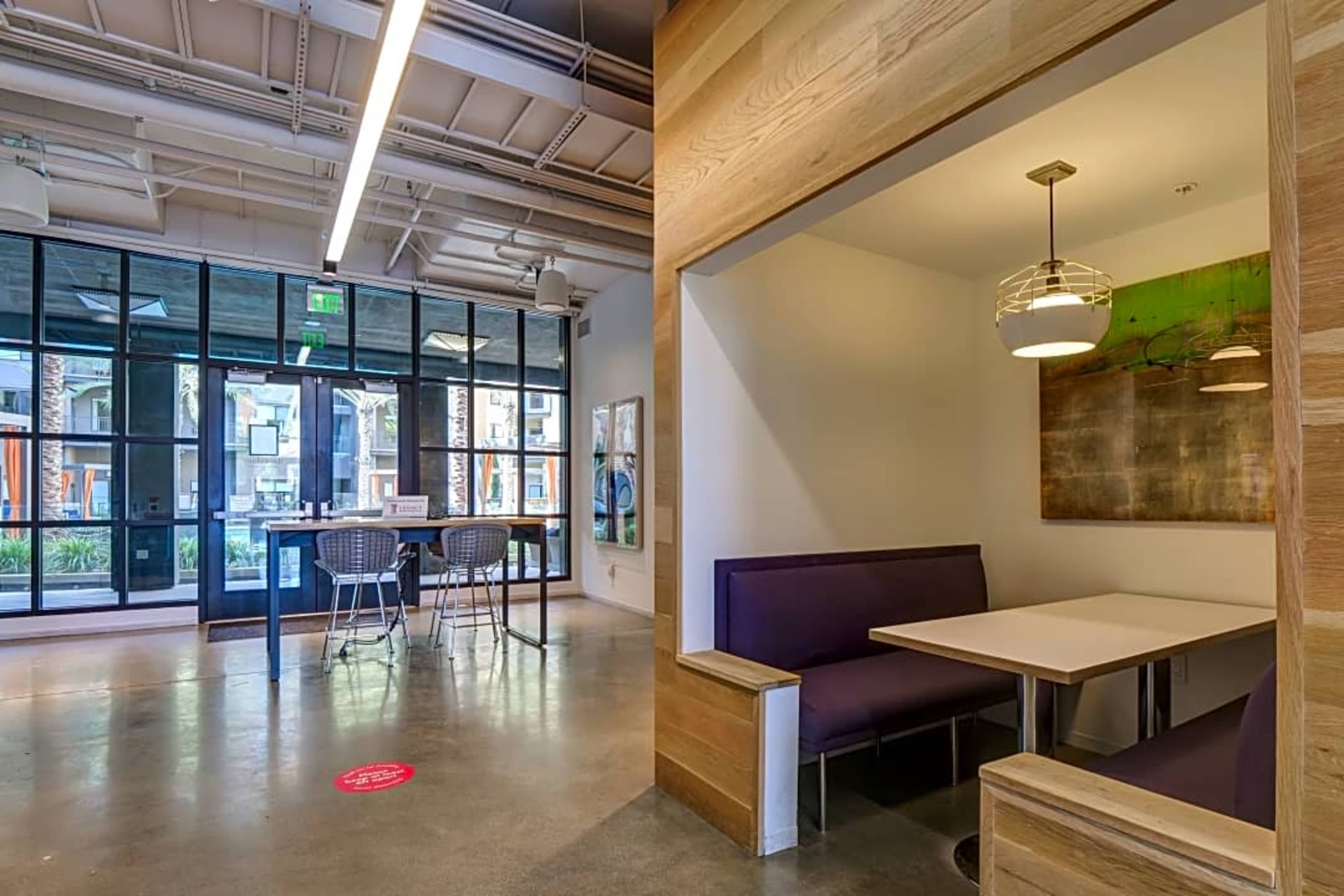 A modern interior space featuring a seating booth and a communal table under bright lighting, with large windows overlooking an outdoor area.