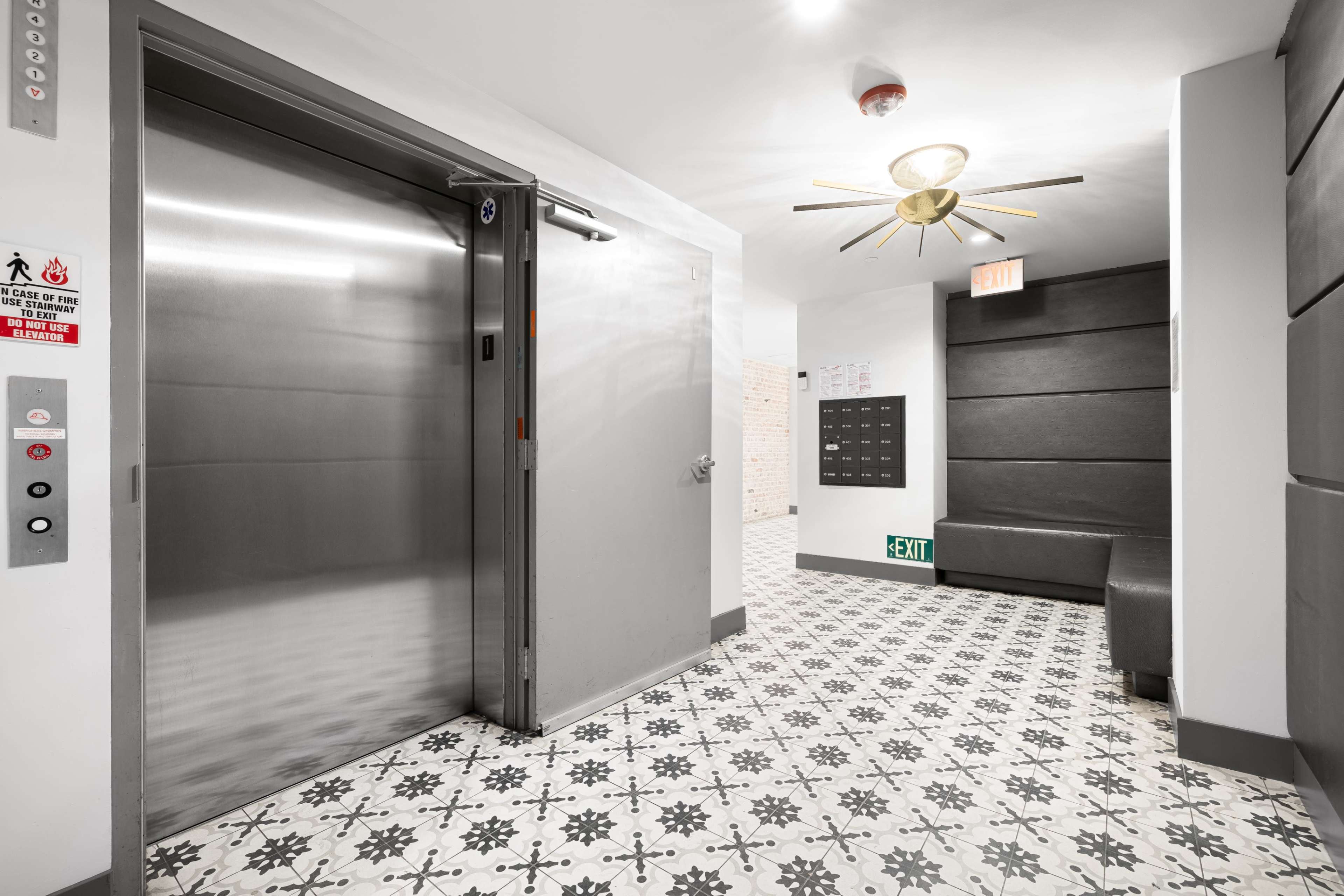 The image shows a hallway with a lift on the left, a decorative ceiling light, a bench on the right, and a visible exit sign at the far end.