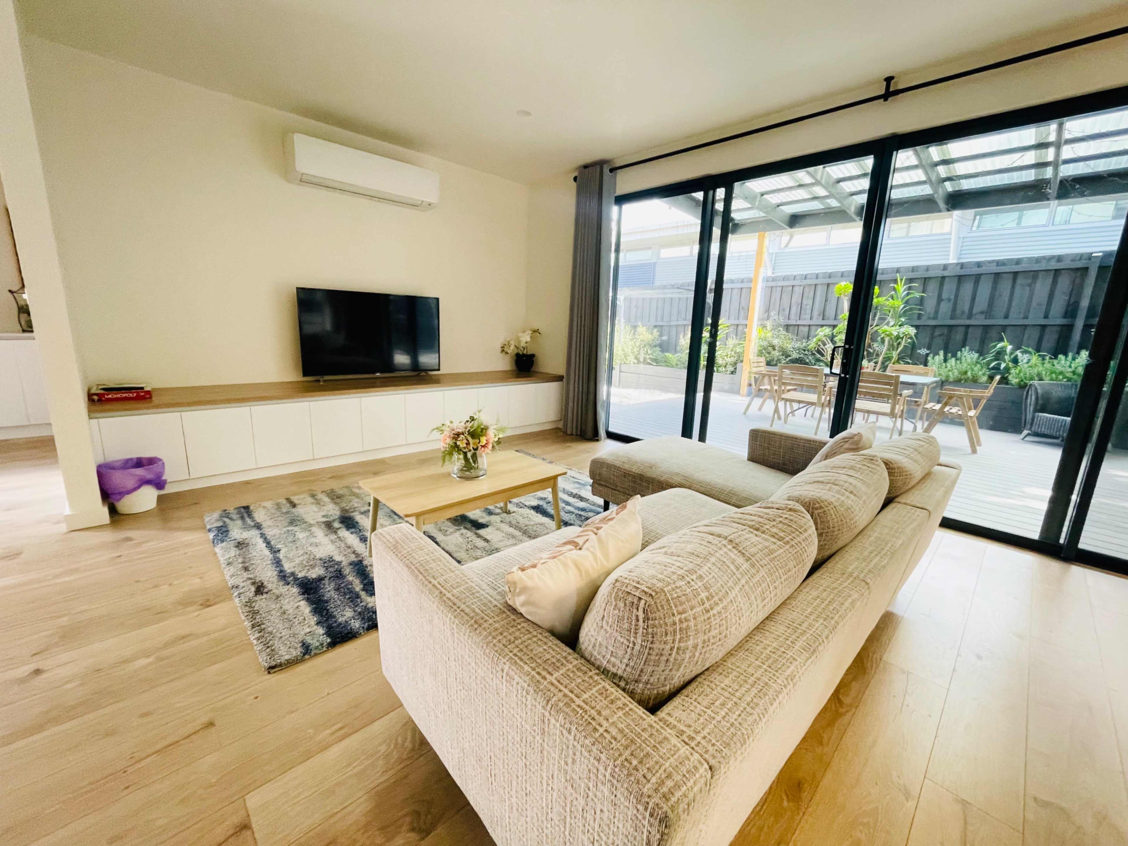 A cozy living room features a sofa facing a wall-mounted television, with large glass doors leading to an outdoor deck surrounded by greenery.