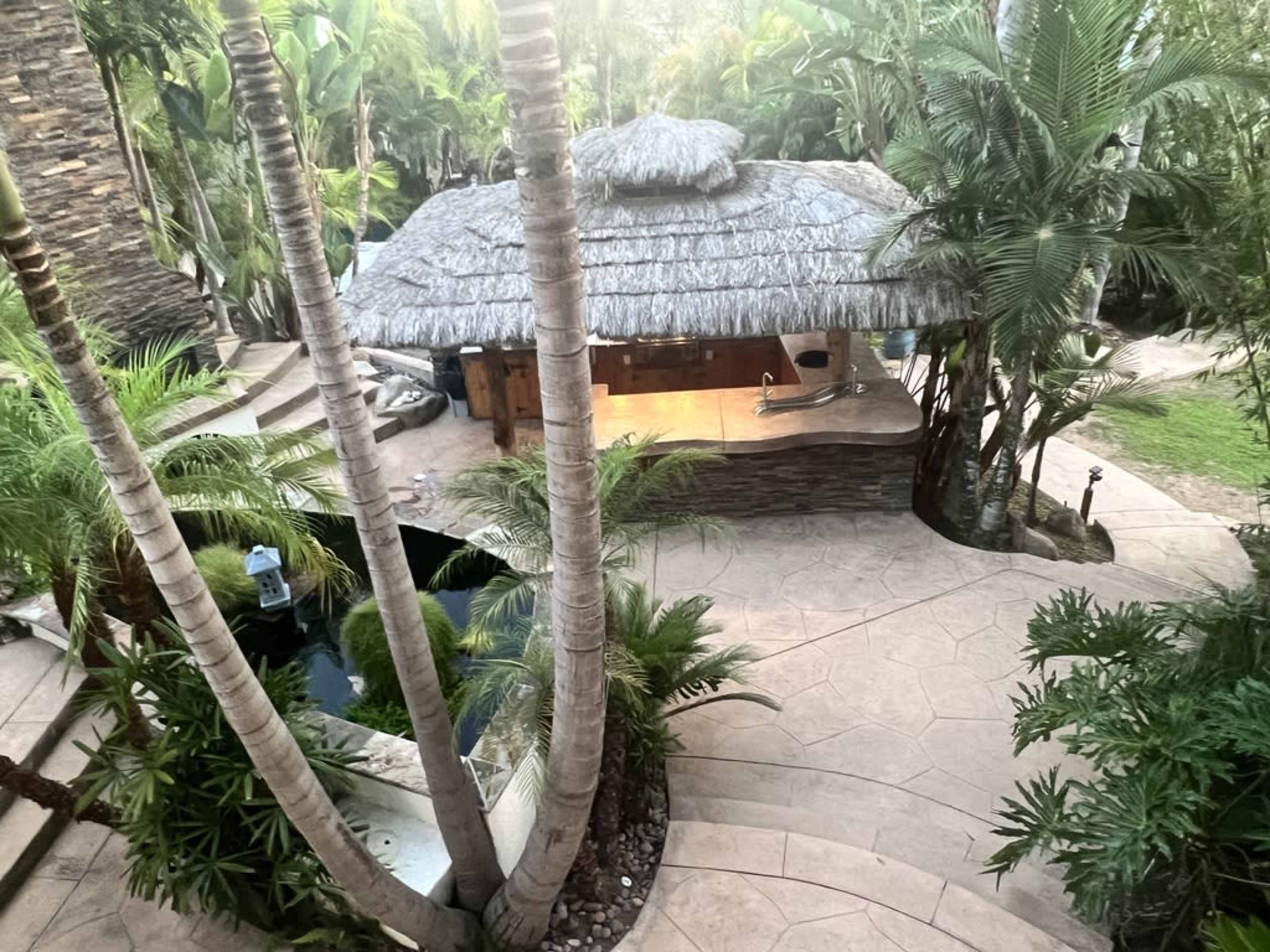 The image shows a thatched-roof hut surrounded by tropical greenery and a water feature, with stone pathways leading towards it.