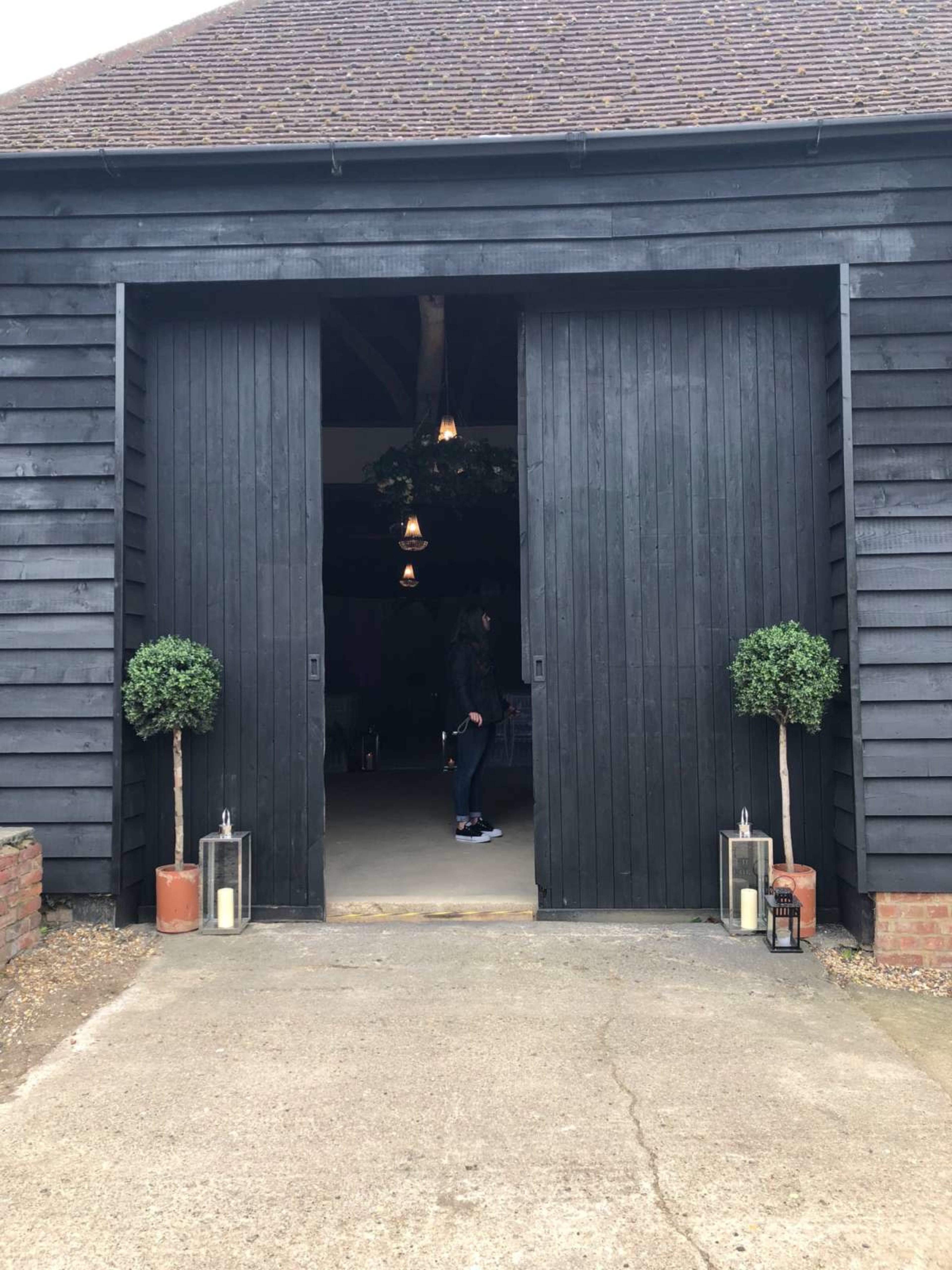 A person walks through large open doors of a dark wooden barn, flanked by potted topiary and lanterns on either side.