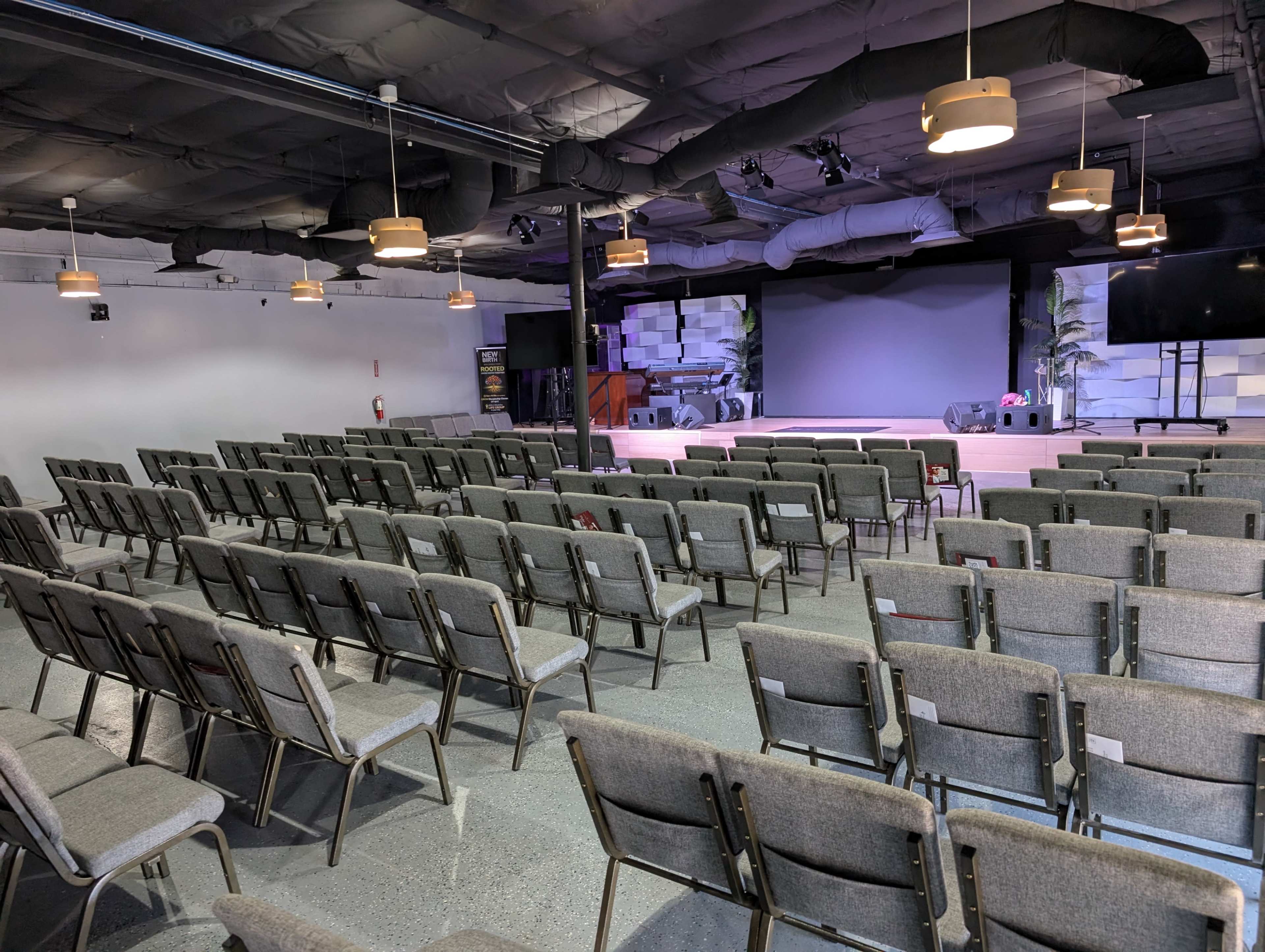 The image shows an empty auditorium with rows of gray chairs facing a stage that features a large screen and lighting fixtures.