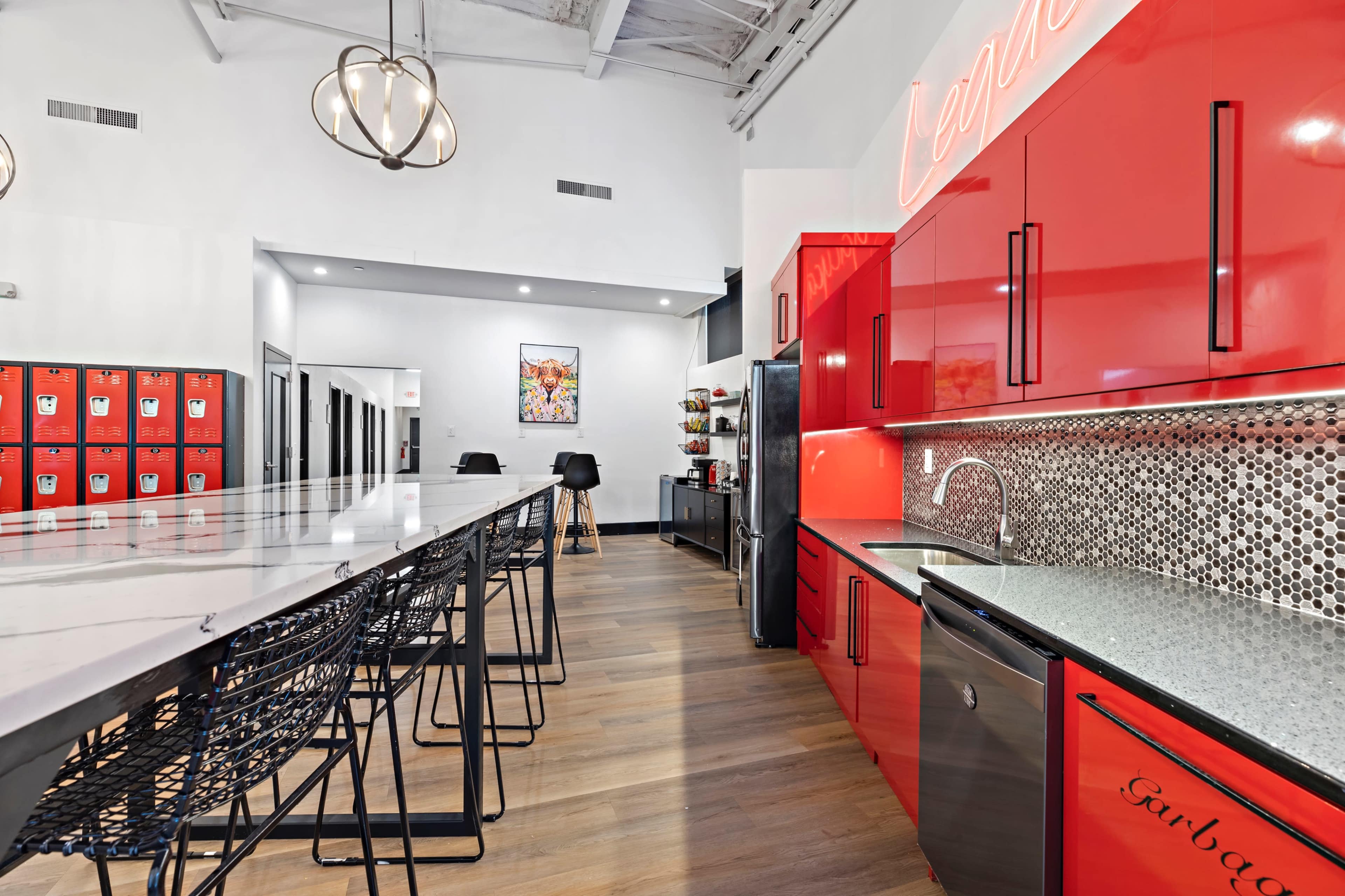 The image shows a modern kitchen with red cabinets and a long countertop, featuring bar stools, a refrigerator, and decorative storage lockers visible in the background.