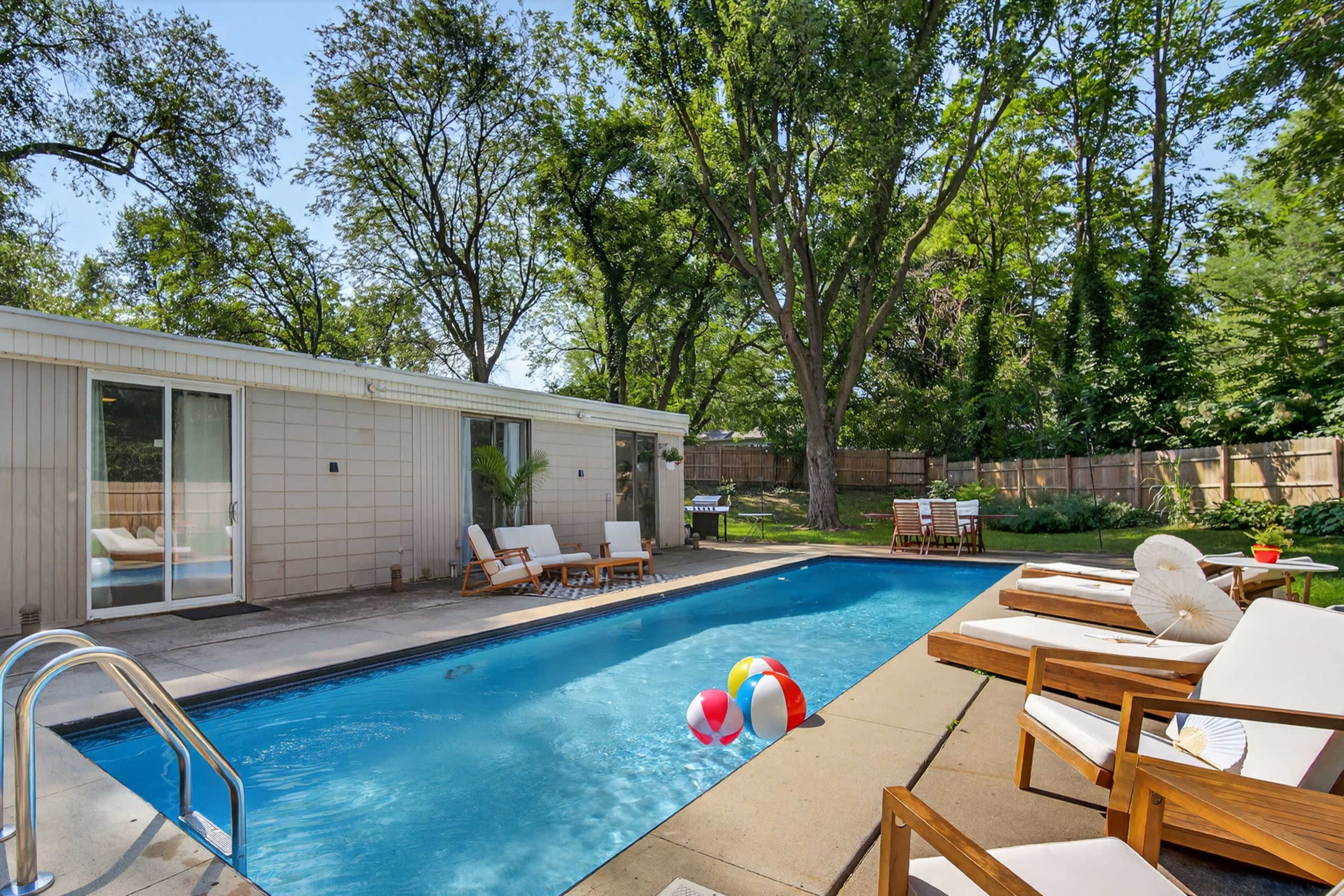 A swimming pool with colorful beach balls is surrounded by wooden lounge chairs and a modern house, all set in a shaded outdoor area with trees.