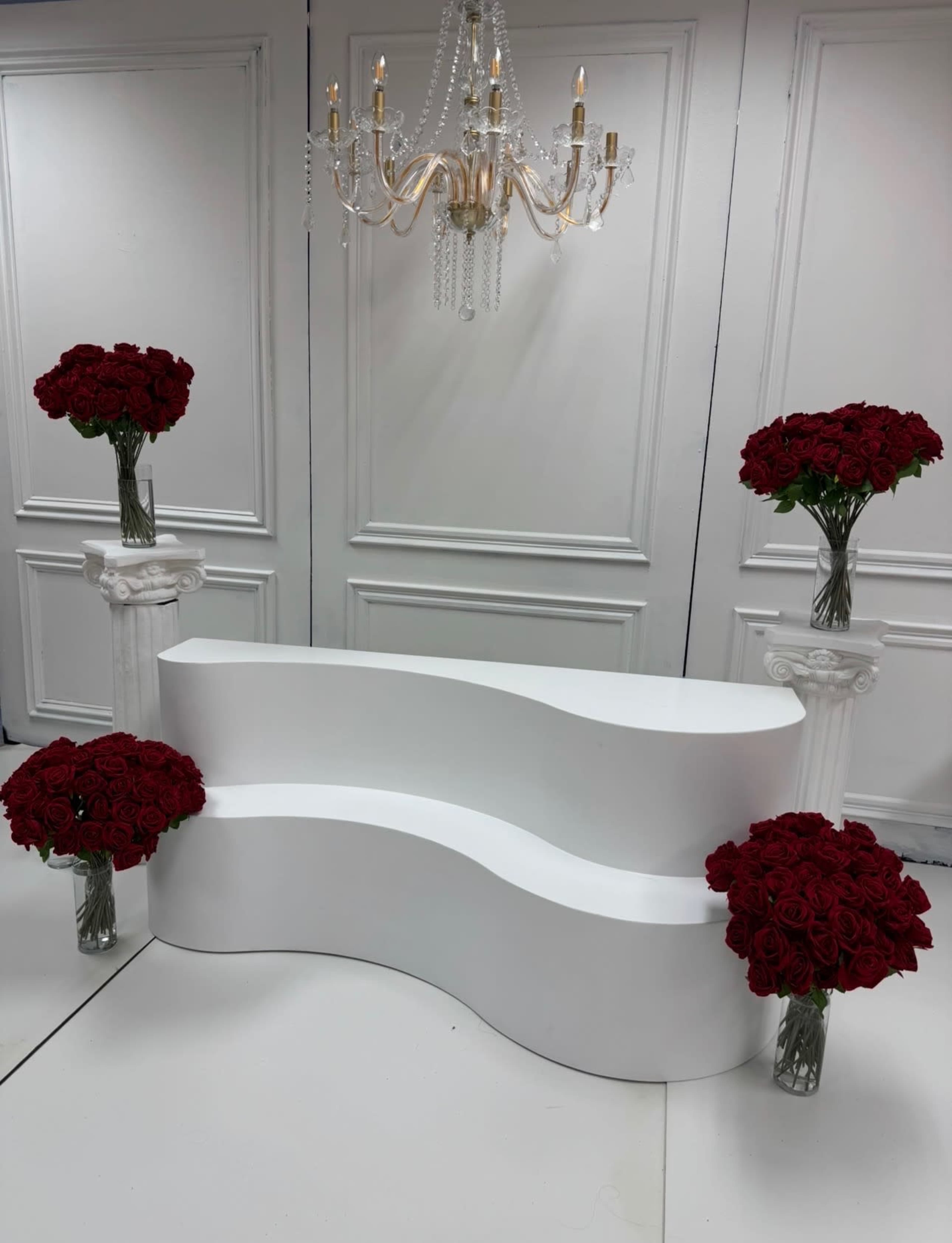 The image shows a white curved bench set in a room with a chandelier overhead, flanked by columns that hold bouquets of red roses.