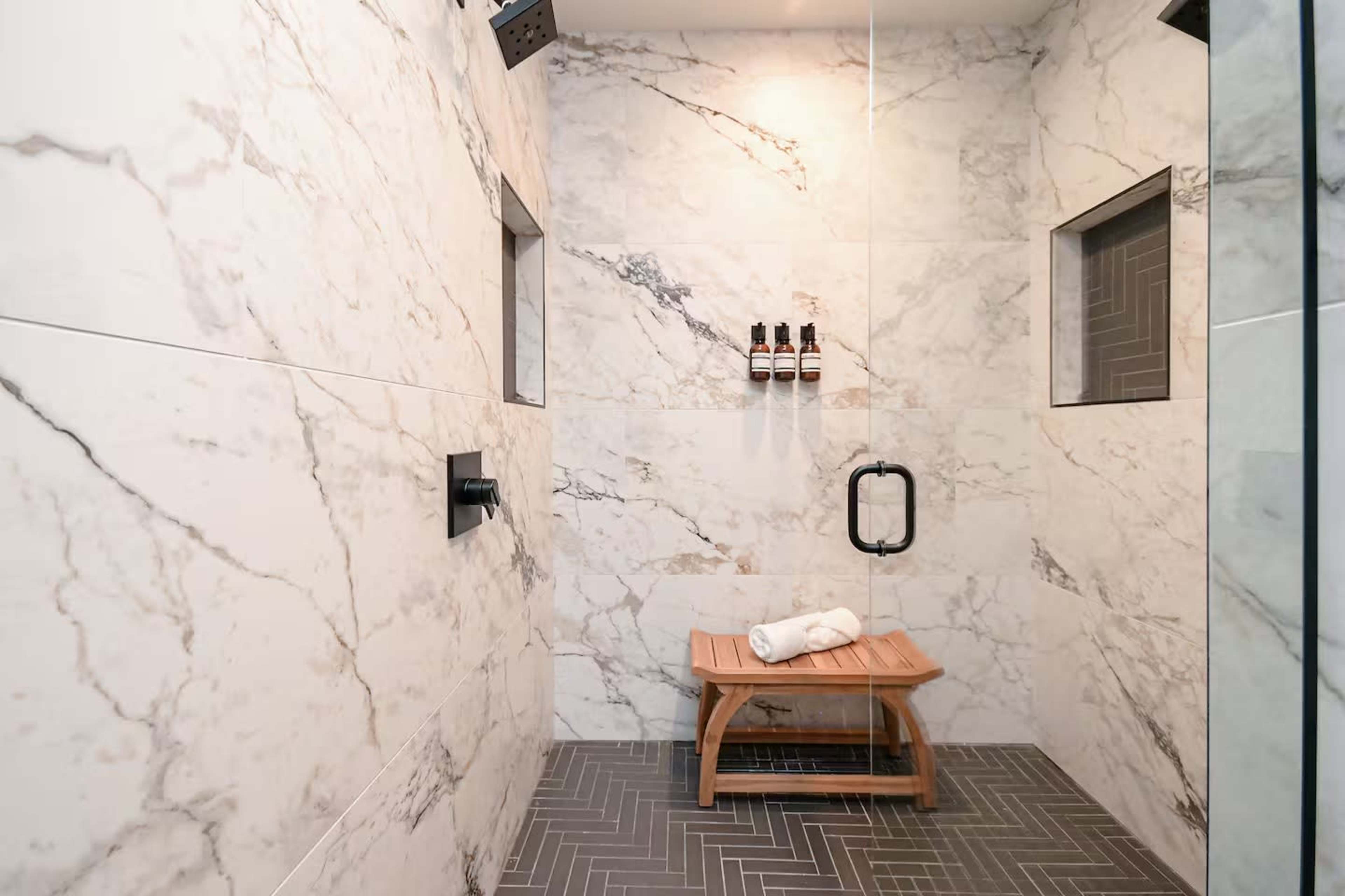 The image shows a modern shower with marble walls, a wooden bench, and three bottles mounted on the wall.