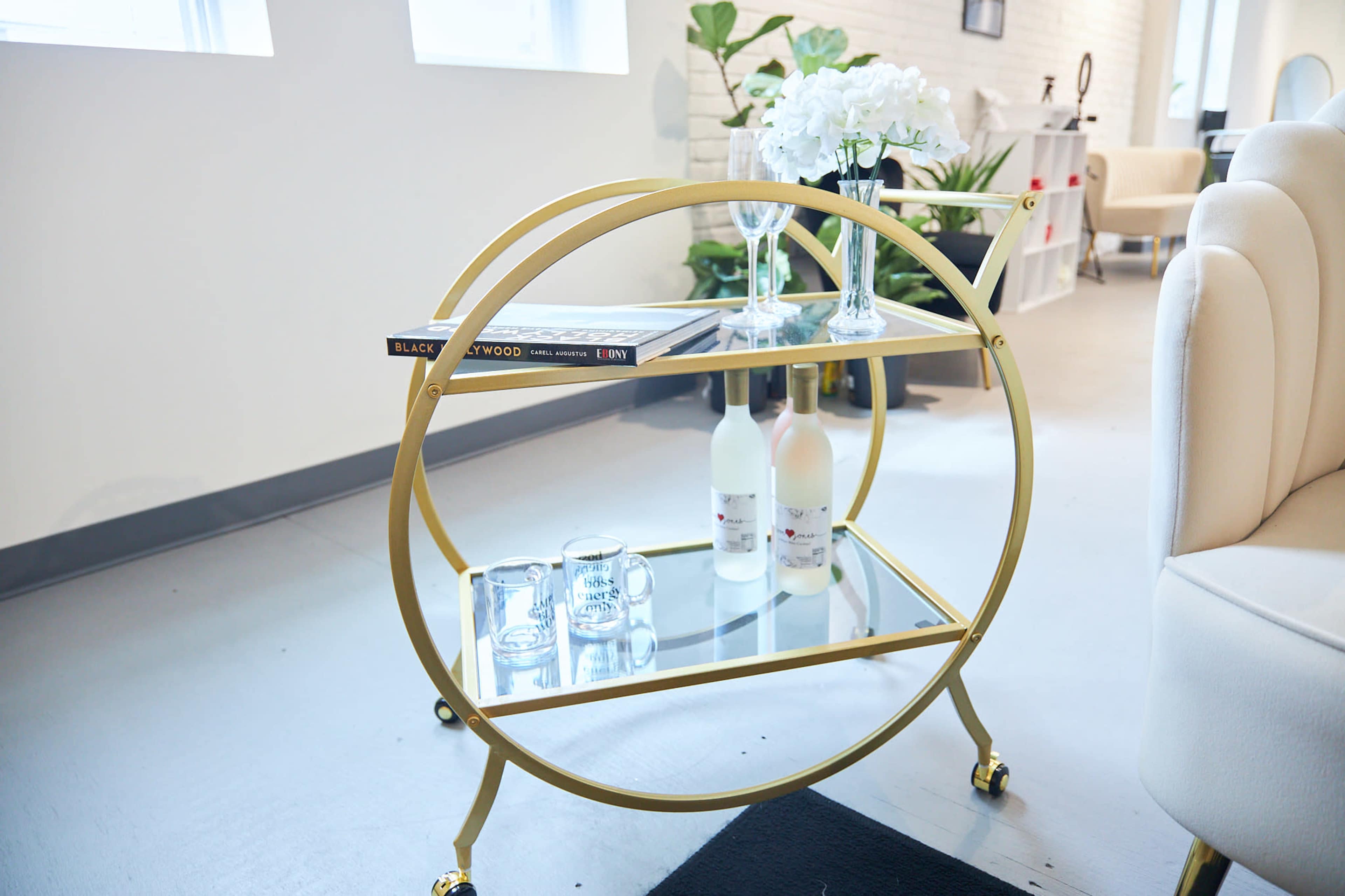 A gold circular bar cart with two glass shelves holds decorative items, including two bottles, glasses, and a book, set against a light-colored backdrop.