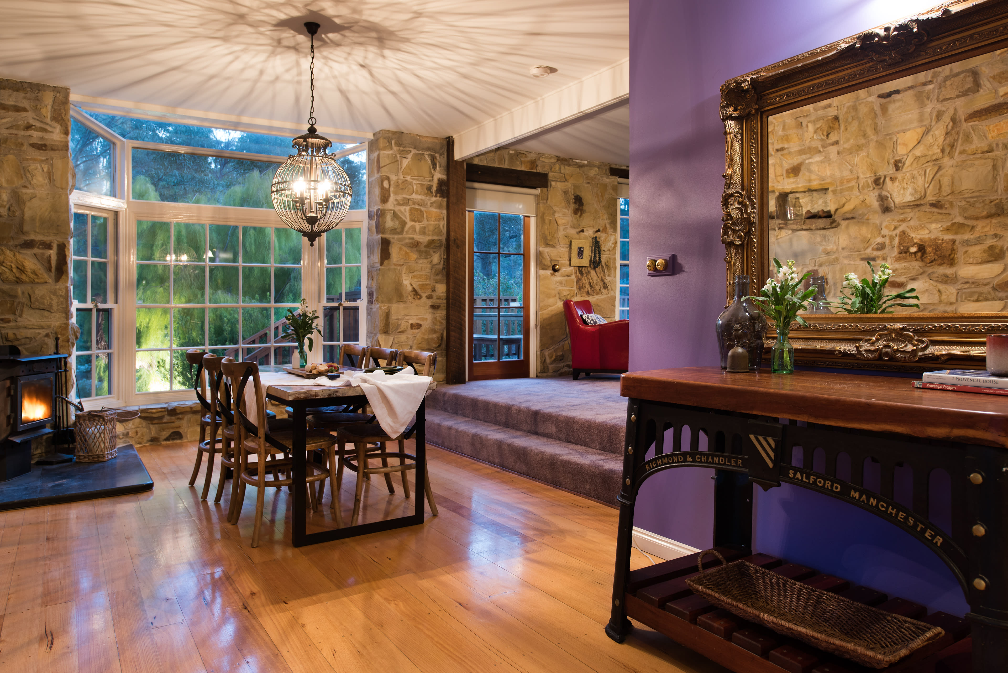 The image shows a cozy dining area with a wooden table and chairs, illuminated by a pendant light, adjacent to a living space featuring a stone wall and a red armchair.