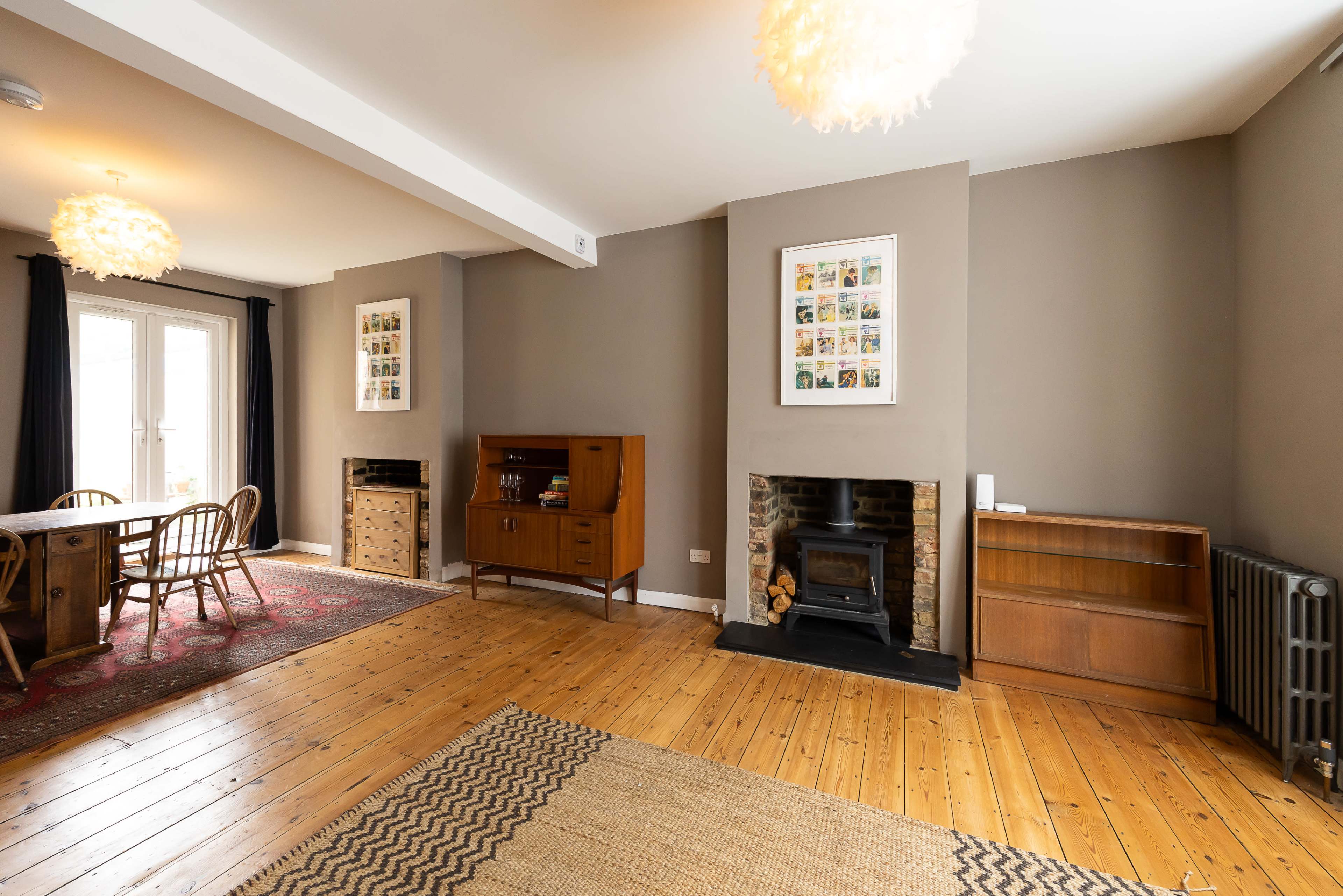 The image shows a spacious living area with wooden flooring, featuring a dining table, an empty bookshelf, and two decorated walls with framed photographs, alongside a fireplace.