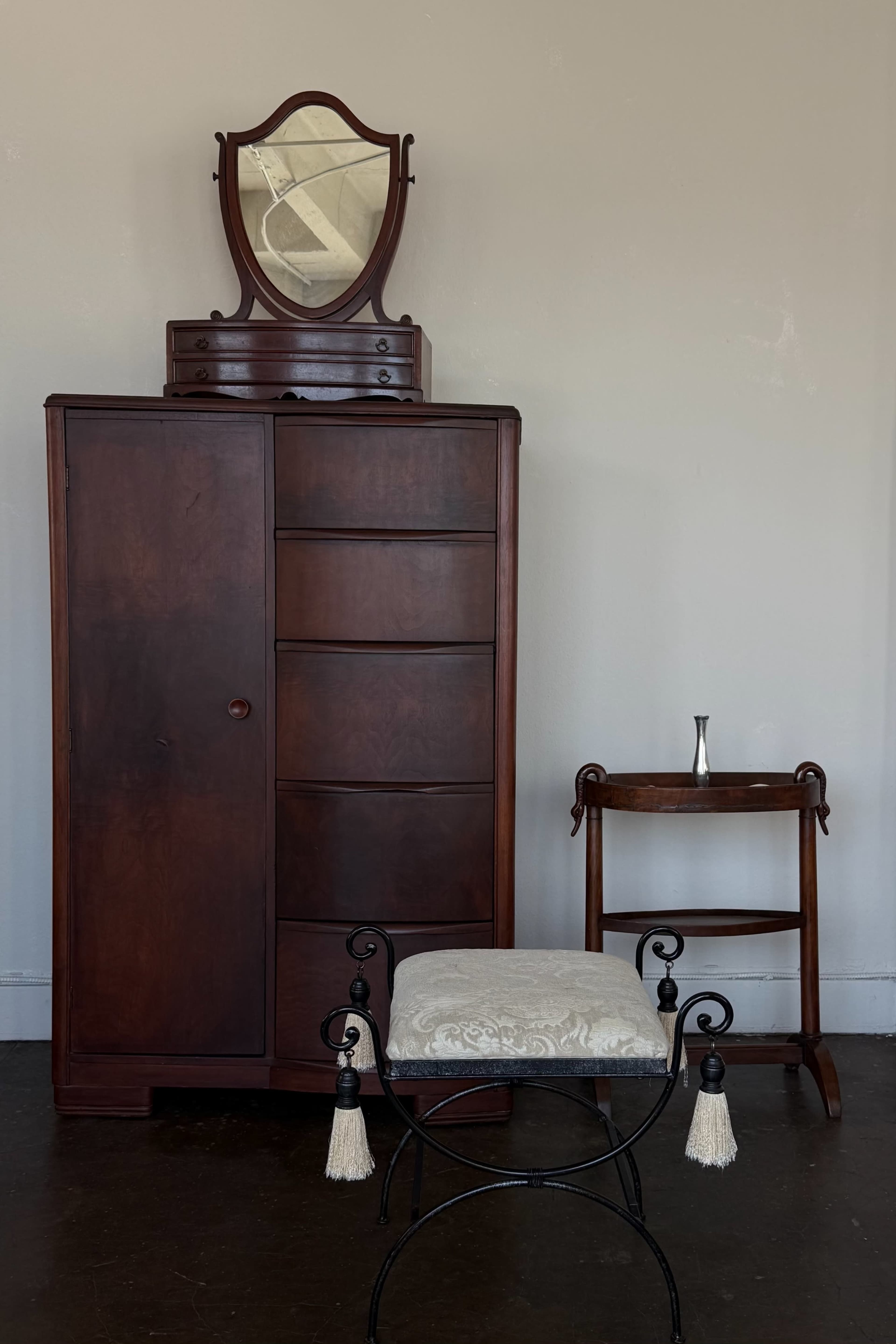 The image shows a dark wooden dresser with a mirror on top, accompanied by a vintage folding stool and a small stand holding a vase.