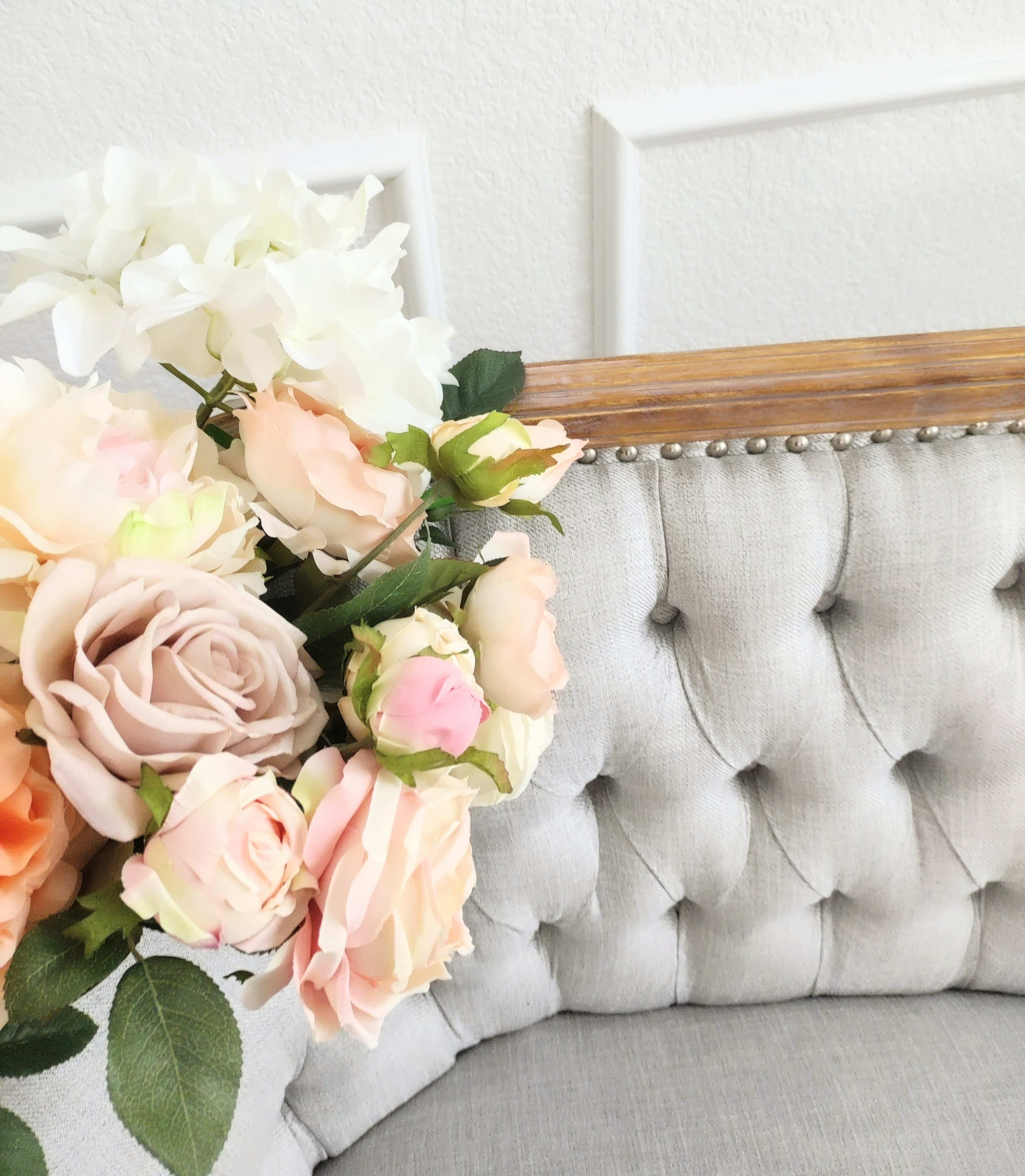 A bouquet of artificial roses and white flowers is positioned on a tufted gray fabric cushion with a wooden frame in the background.