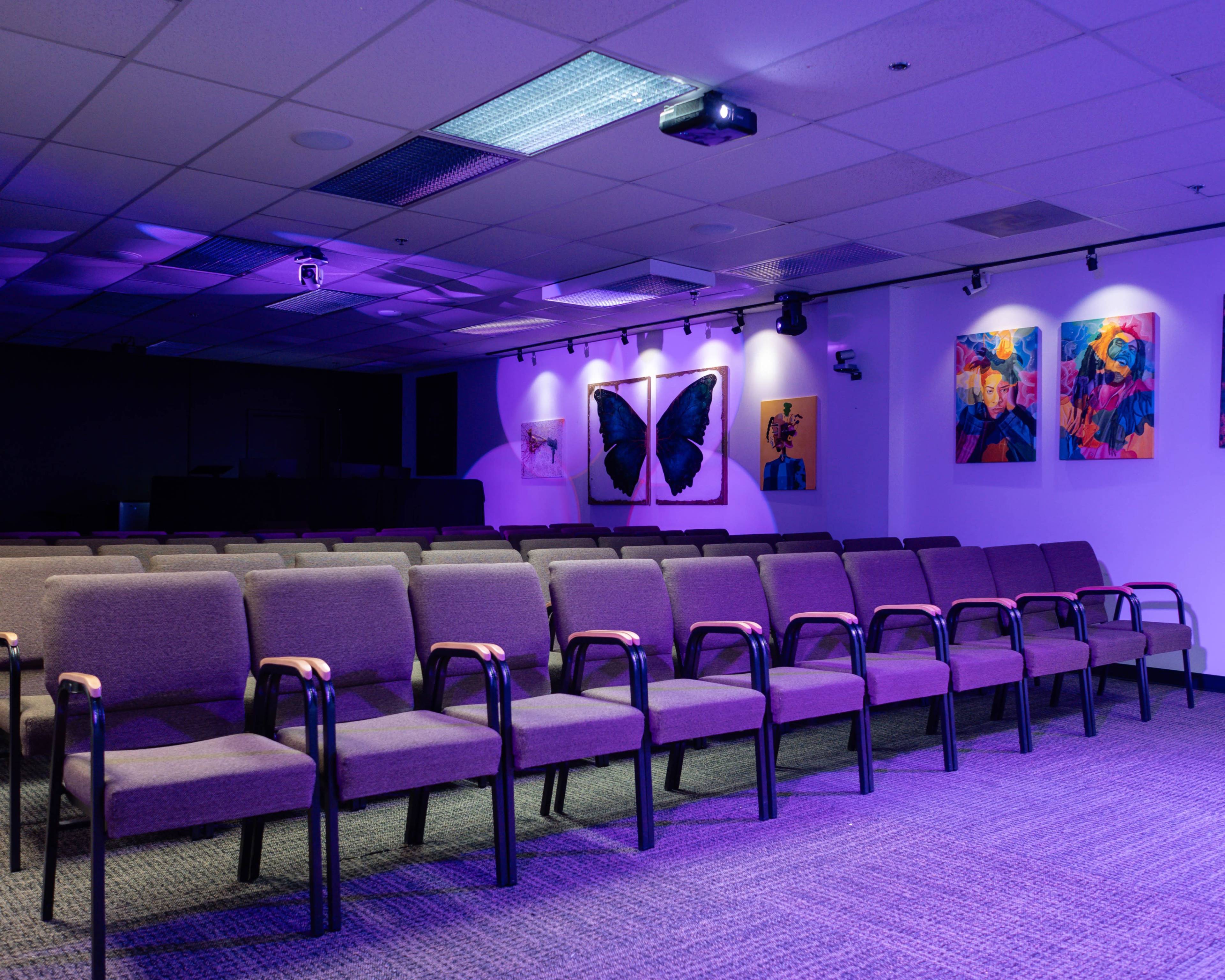 A row of empty chairs is arranged neatly in a dimly lit room featuring colorful butterfly and abstract paintings on the walls.