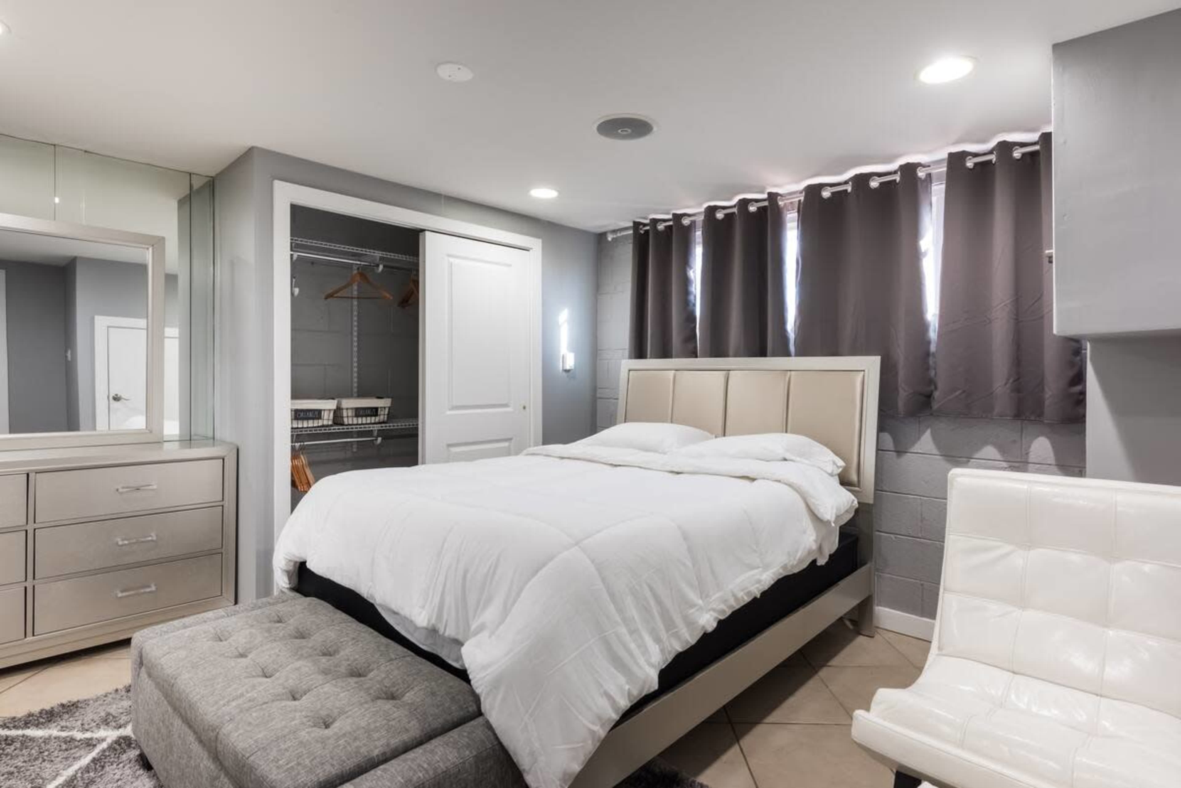 The image shows a modern bedroom featuring a bed with white bedding, a gray accent chair, a mirrored dresser, and dark gray curtains.