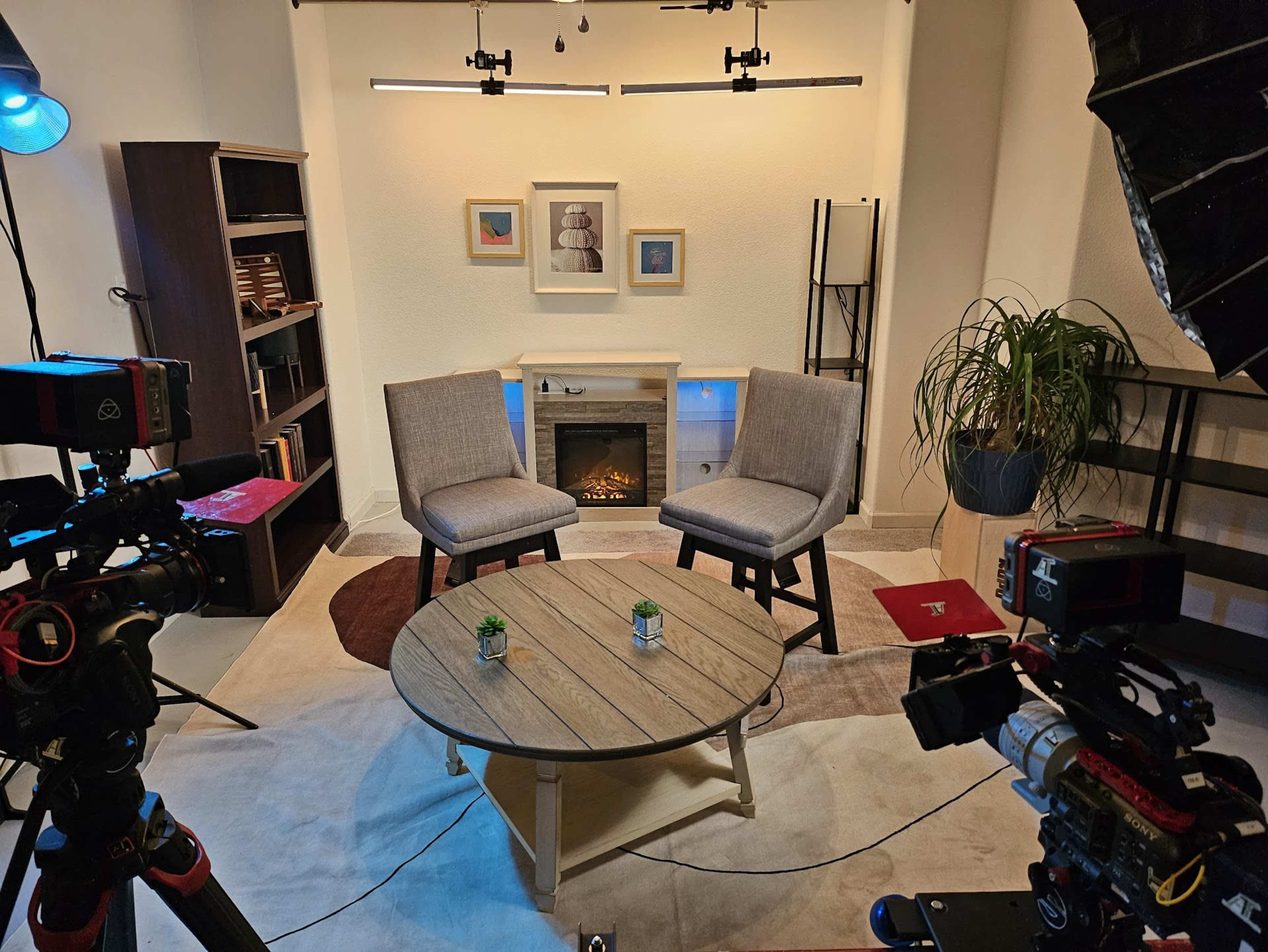 The image shows a modern, well-lit studio setup featuring two gray chairs facing a coffee table, with a faux fireplace and bookshelf in the background.