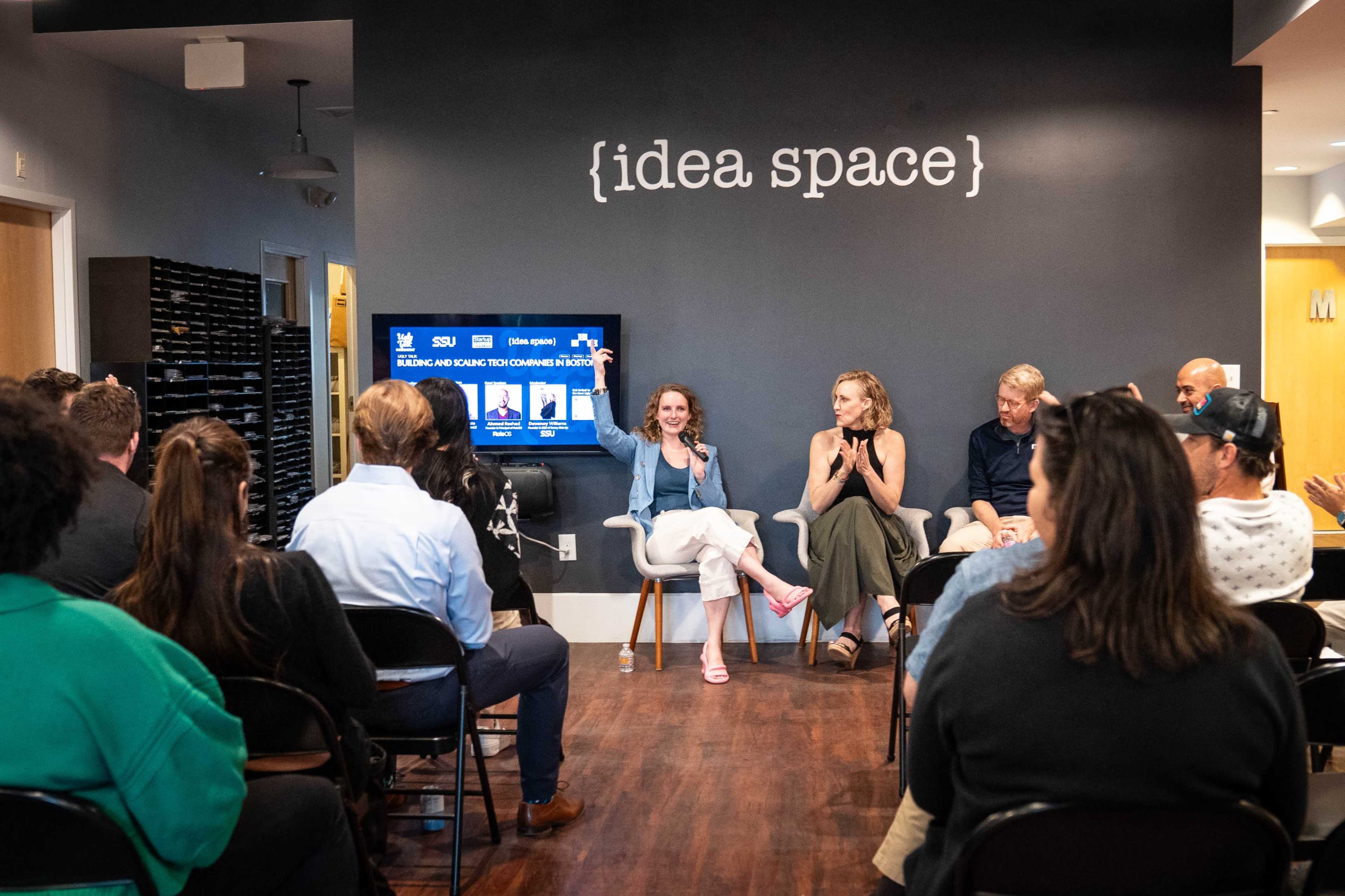 A panel discussion is taking place in a modern workspace, with audience members seated in front of speakers discussing various topics, while a screen displays information related to the event.