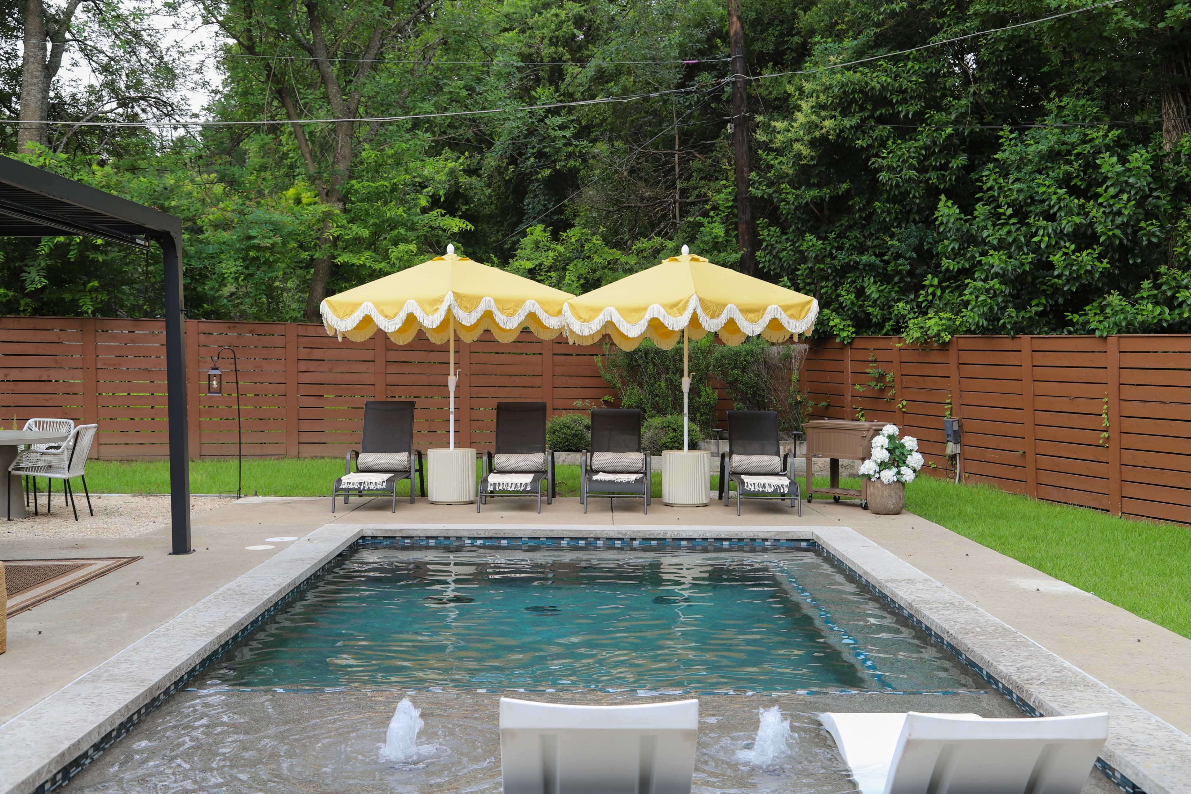 A backyard scene features a swimming pool surrounded by lounge chairs and two yellow umbrellas, with a wooden fence and greenery in the background.