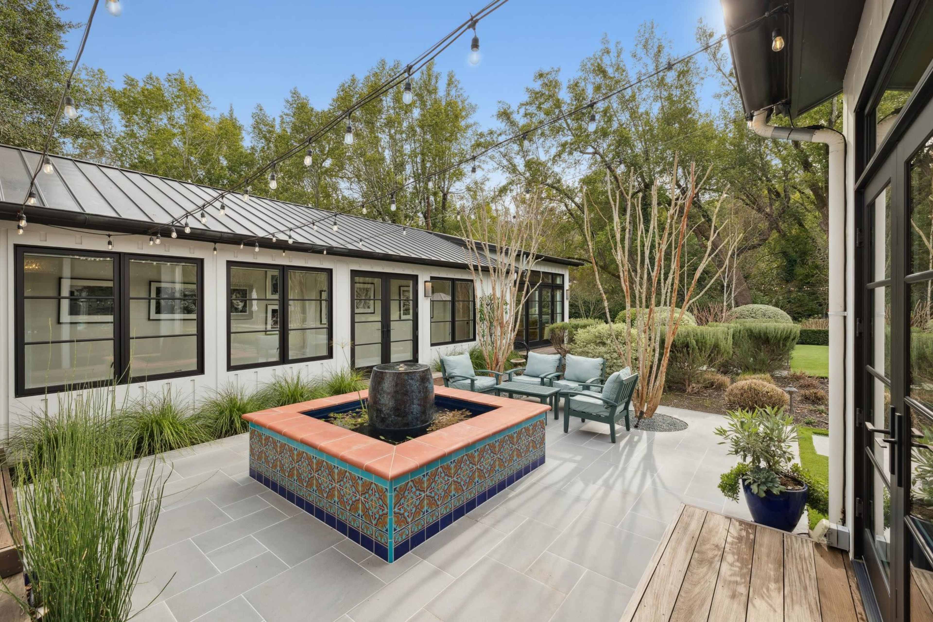 The image shows a courtyard featuring a tiled fountain surrounded by seating and greenery, with a modern building in the background.