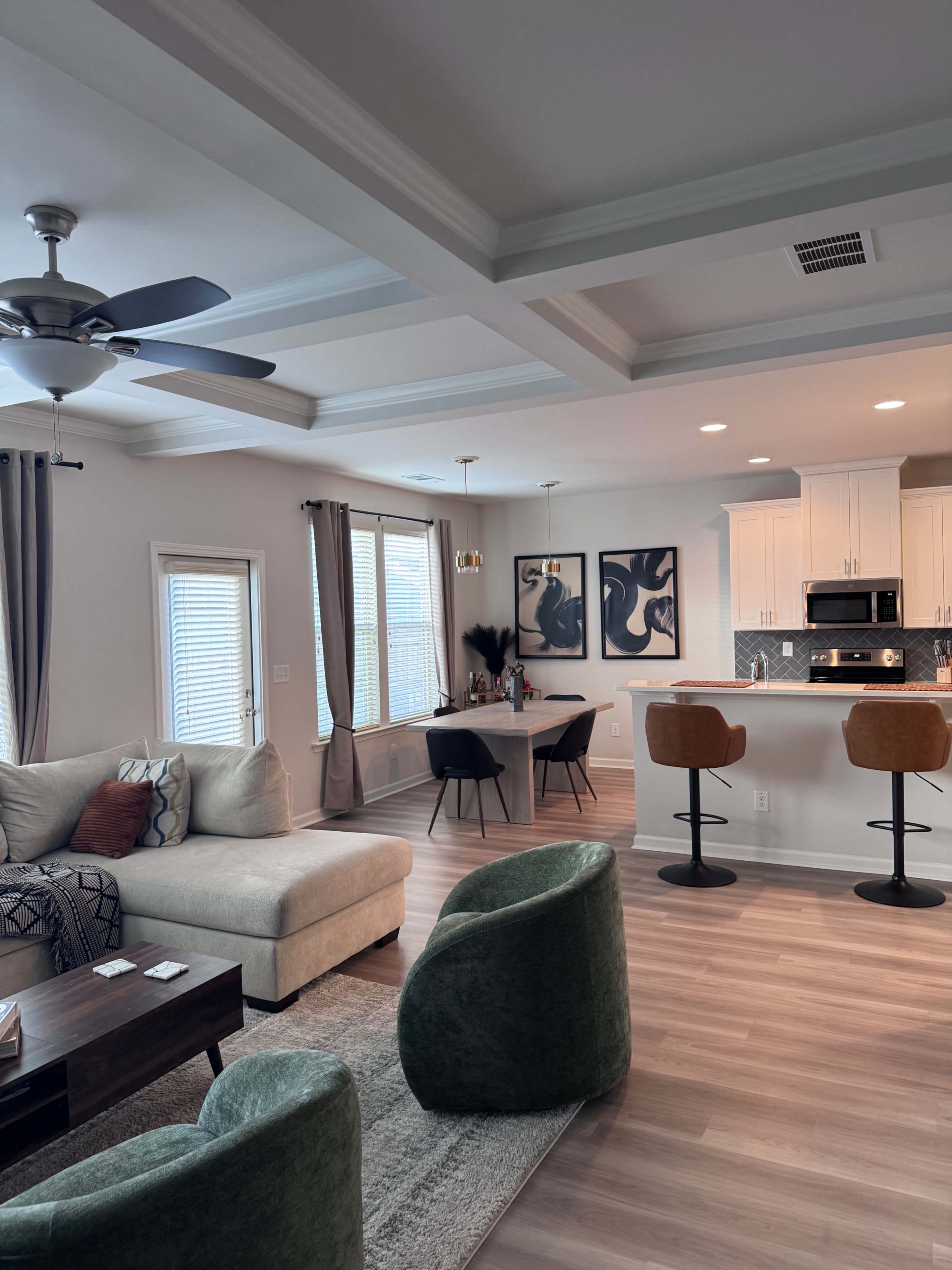 The image shows a modern living space featuring a sectional sofa, a dining table, and a kitchen with bar stools, all illuminated by natural light from the windows.