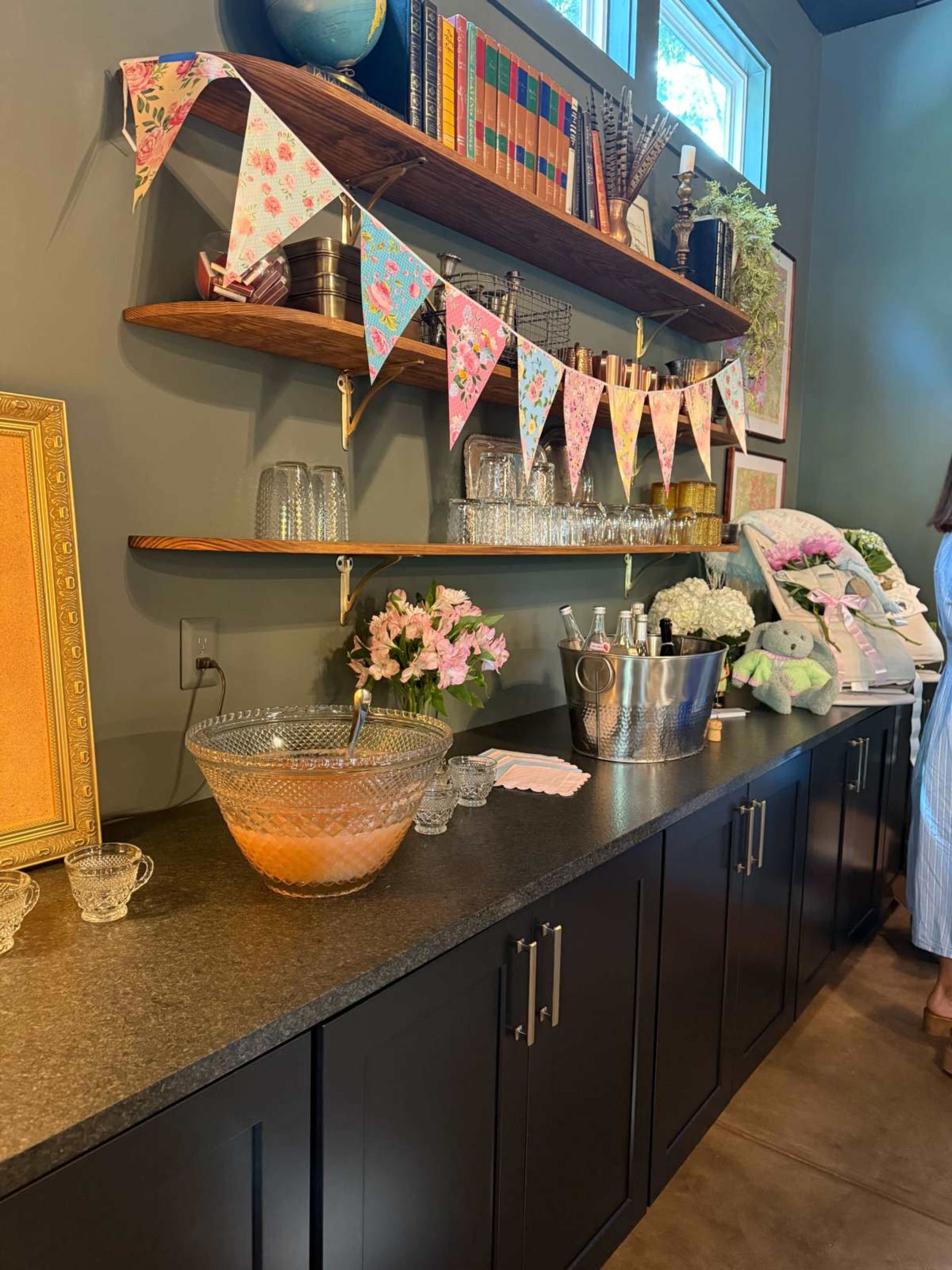 The image shows a decorative table setup featuring a punch bowl, glassware, flowers, and a banner on a dark countertop beneath wall-mounted shelves with books and plants.