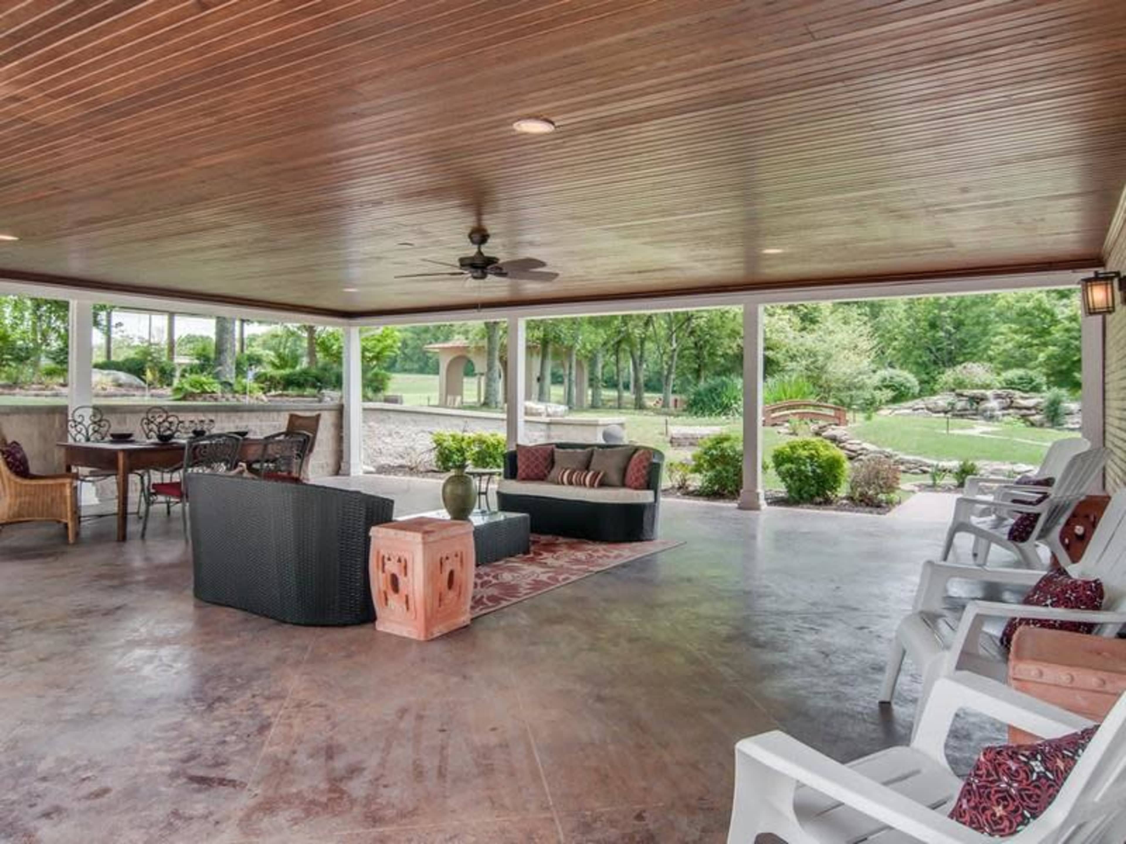 The image shows a spacious covered patio with various seating arrangements, a ceiling fan, and views of a landscaped garden.