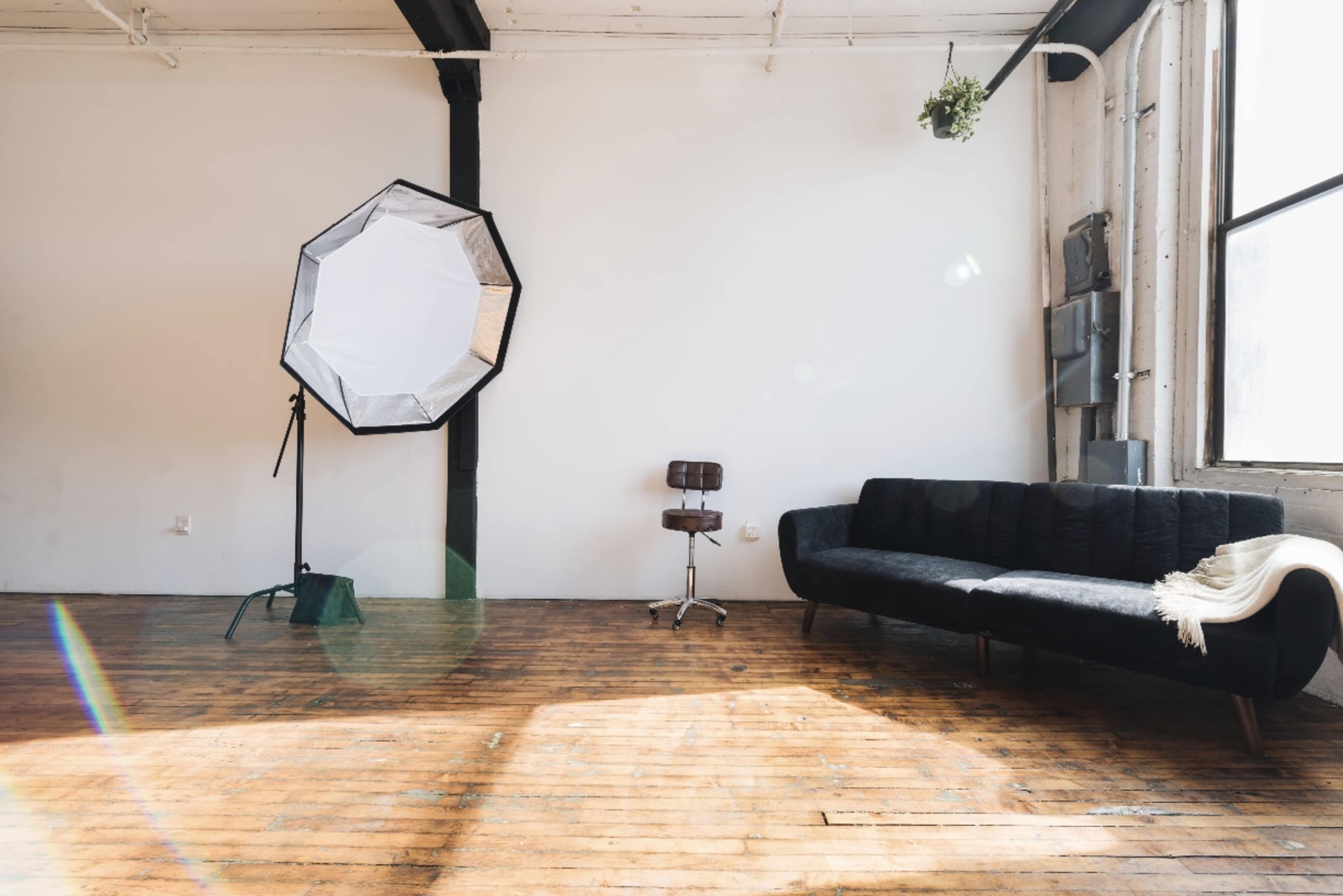 A bright, spacious room with a black sofa, a wooden floor, a photography softbox, and a metallic stool.